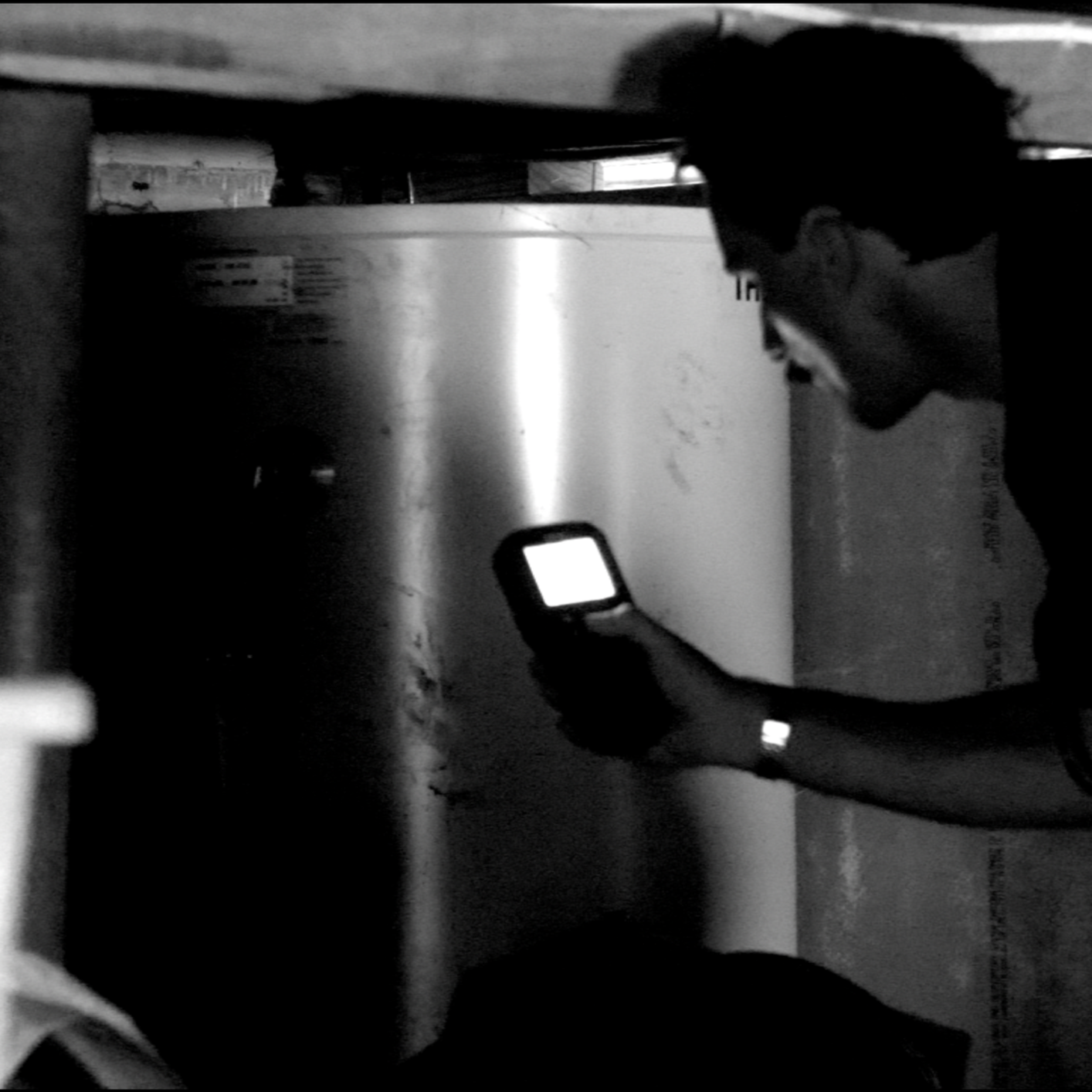 A person with glasses and short hair looking at a phone, standing near a large metallic object in a dimly lit space.