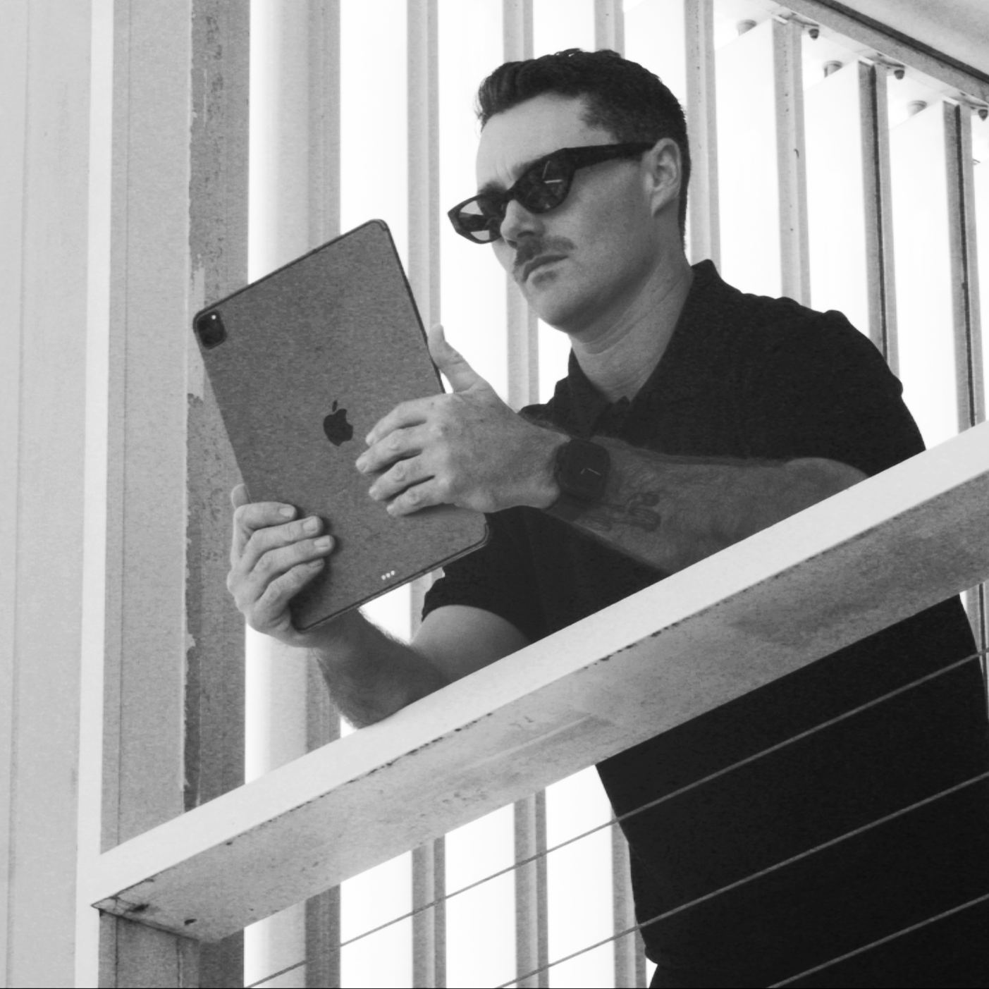 A man wearing sunglasses and a watch, holding an iPad, standing behind a railing.