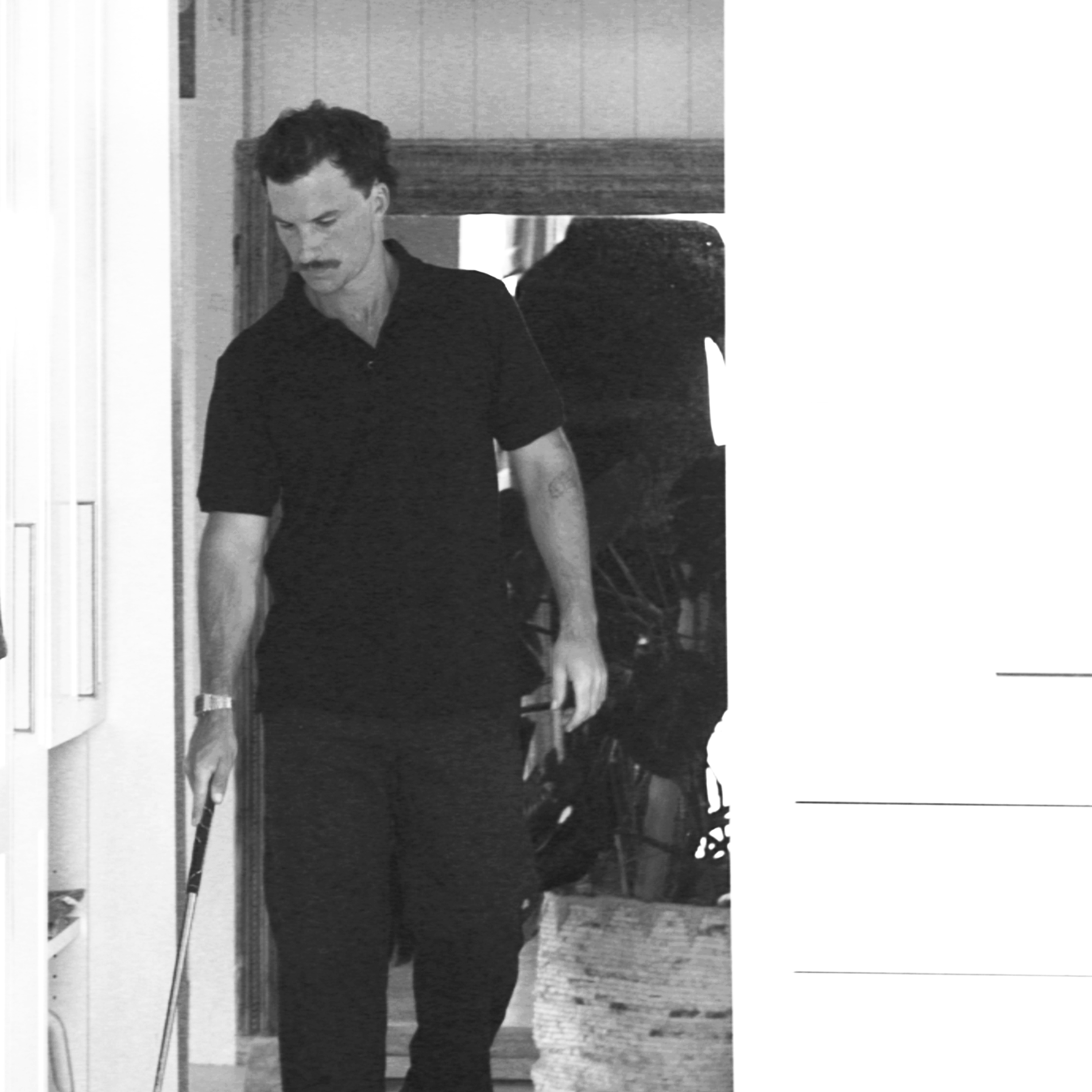 A black and white photo of a man with a mustache using a tapper, wandering through a room with wooden walls and a white dresser.