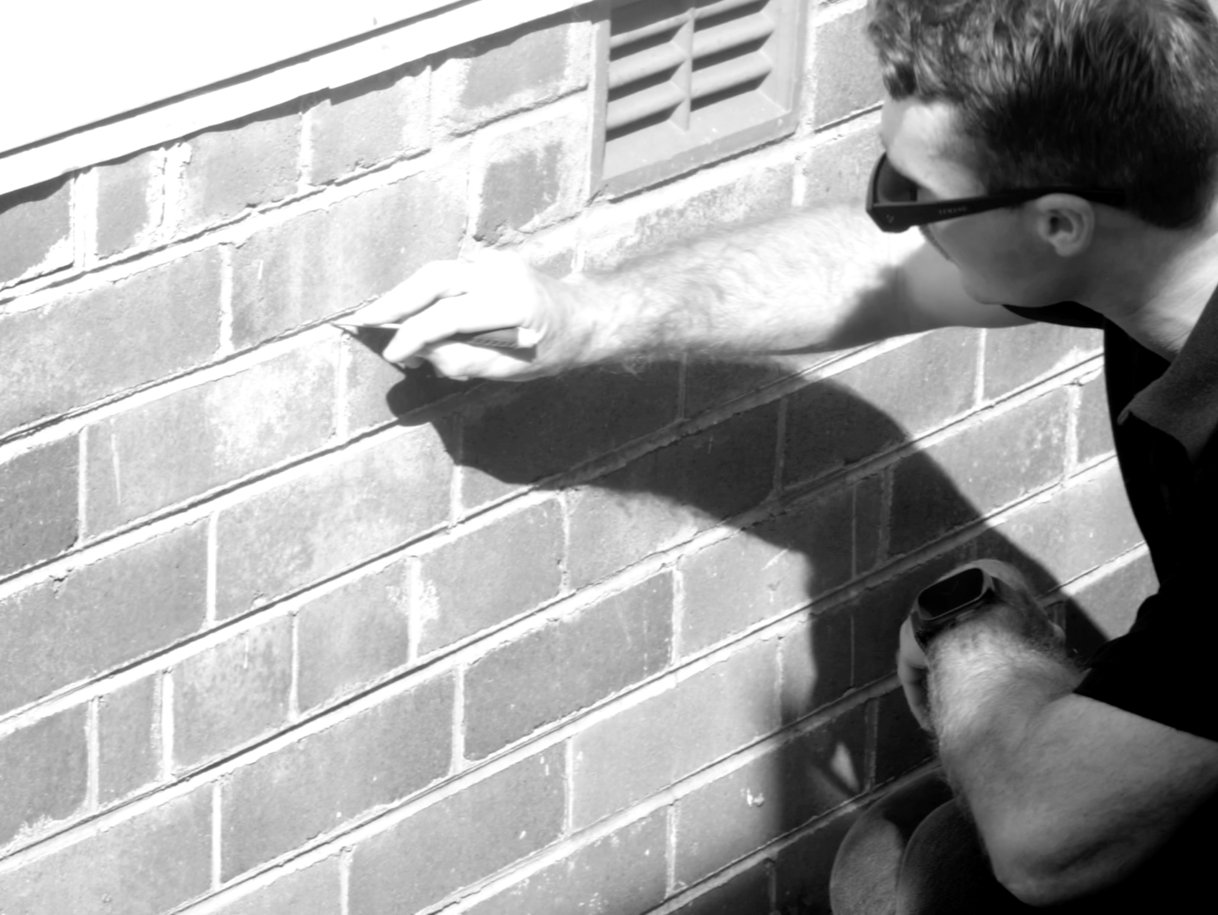 A person sketching or drawing on a brick wall with chalk or a marker. The person is wearing glasses and has short dark hair.