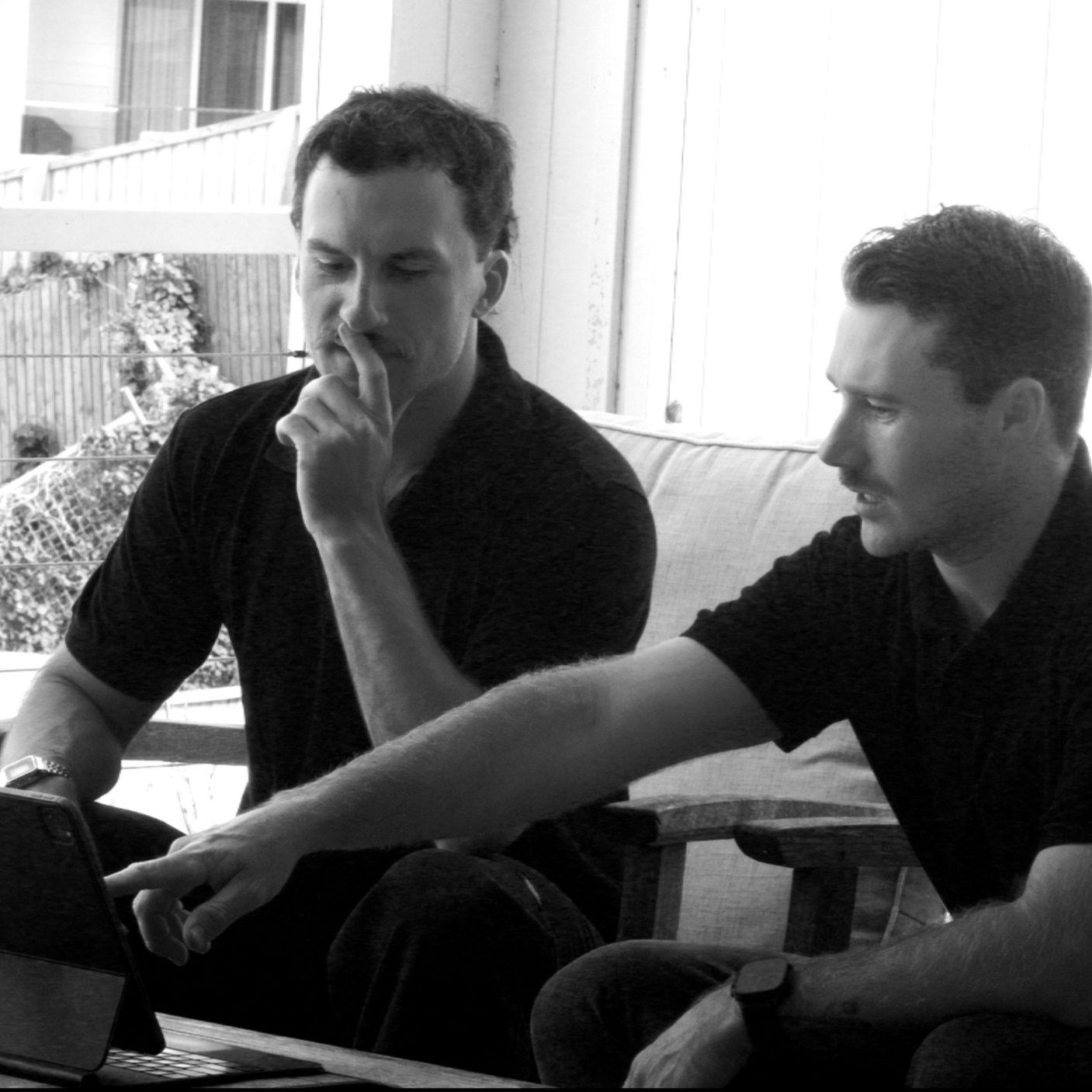Two young men sitting on a couch, looking at a laptop screen, with one pointing at the laptop while the other appears to be thinking.