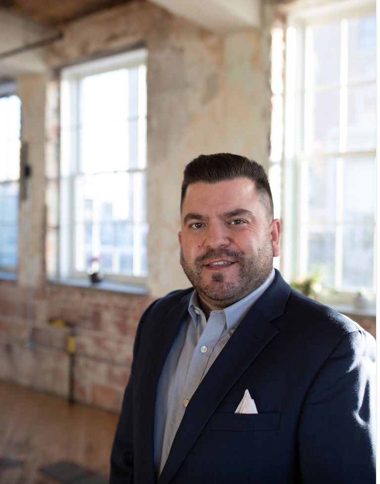A man in a dark suit jacket and light blue shirt standing in a bright, industrial-style room with exposed brick walls and large windows.