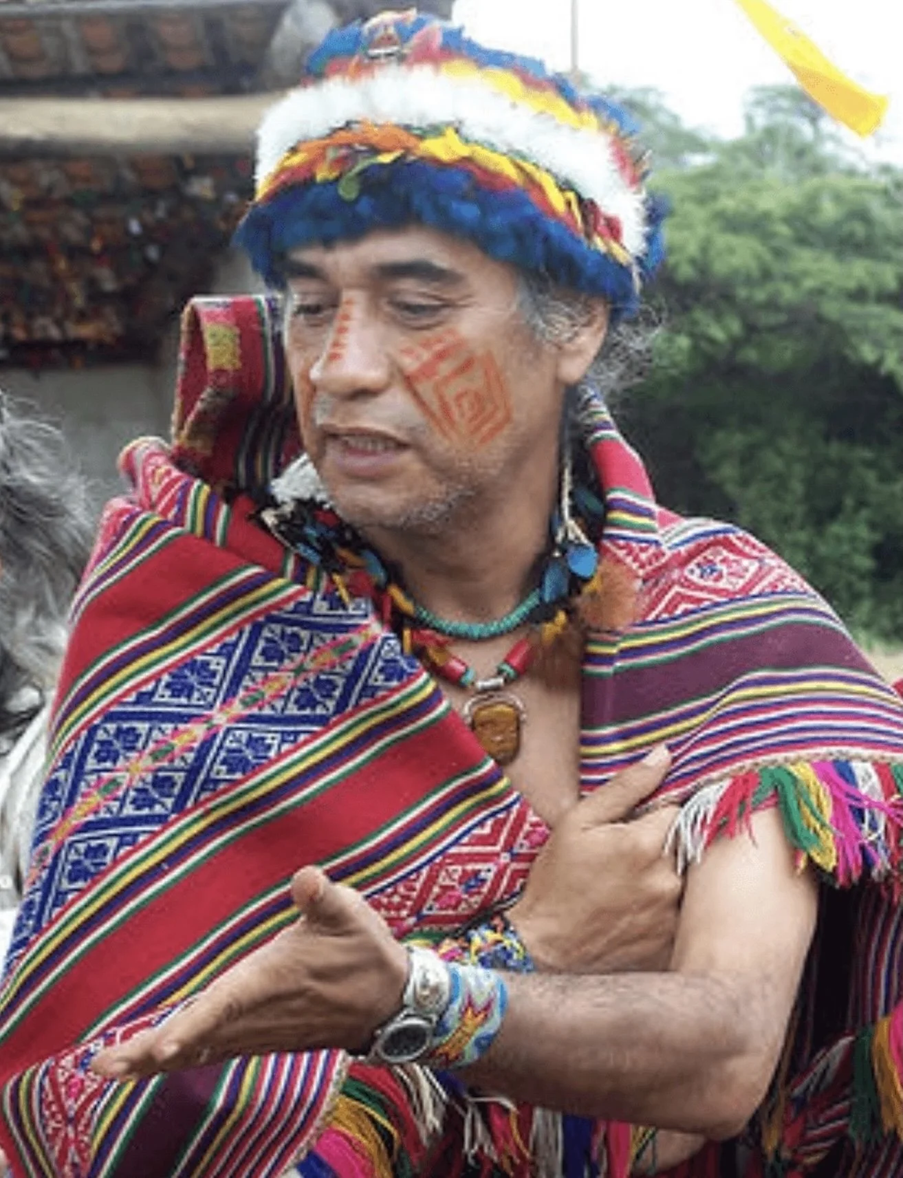 A person wearing colorful traditional indigenous clothing and face paint, embracing another person outdoors.