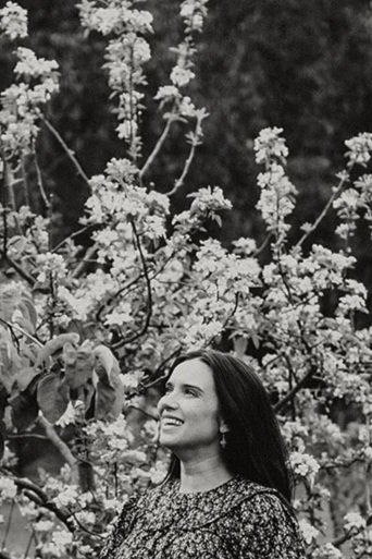 A woman with long dark hair smiling outdoors surrounded by blooming flowers and trees.