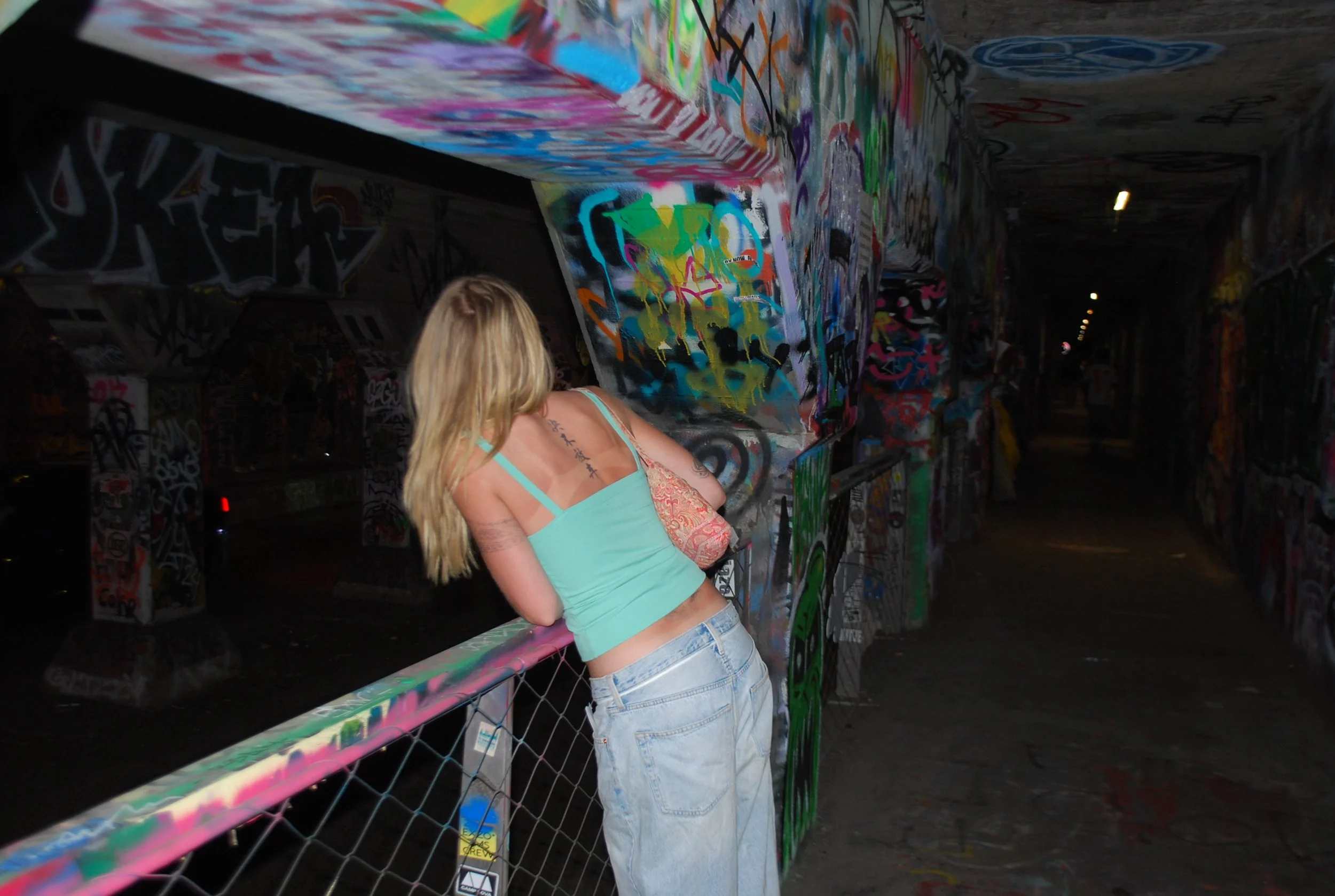 A young woman with long blonde hair in a light blue tank top and jeans leaning on a colorful graffiti-covered bridge rail, looking down in an underground tunnel with graffiti on the walls and ceiling.