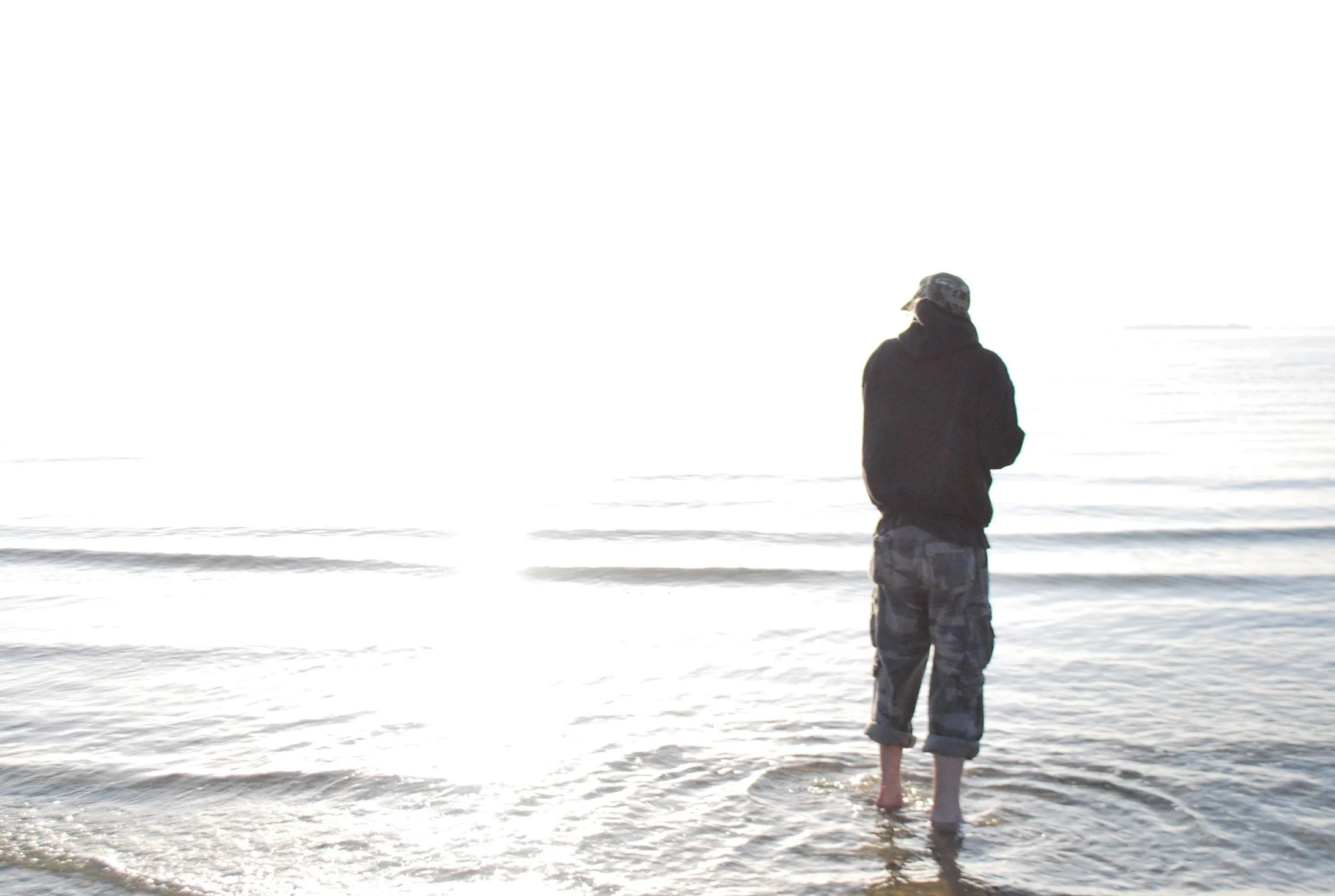 Person standing in shallow water at the beach, facing away, wearing dark clothing and a cap, with the bright ocean and sky in the background.