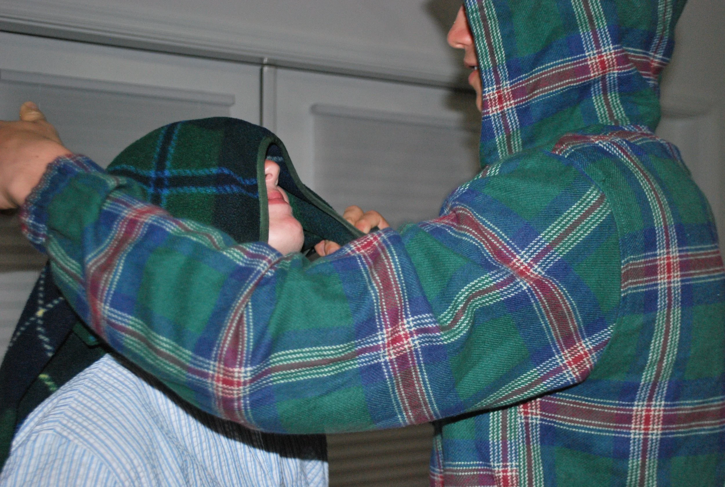 Two individuals in plaid shirts, one of them adjusting the hood of the other's jacket in an indoor setting.