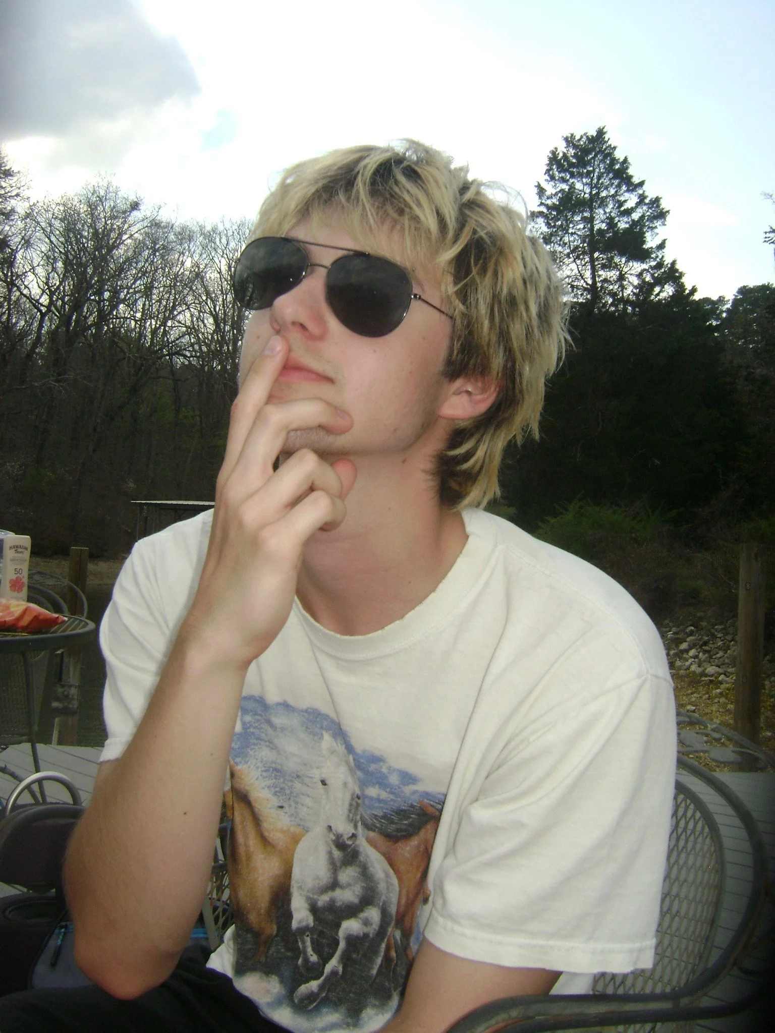 A young man with blonde, slightly wavy hair wearing black sunglasses, a light-colored t-shirt with a graphic of a wolf and a mountain scene, and touching his chin thoughtfully outdoors in a natural setting with trees and cloudy sky in the background.
