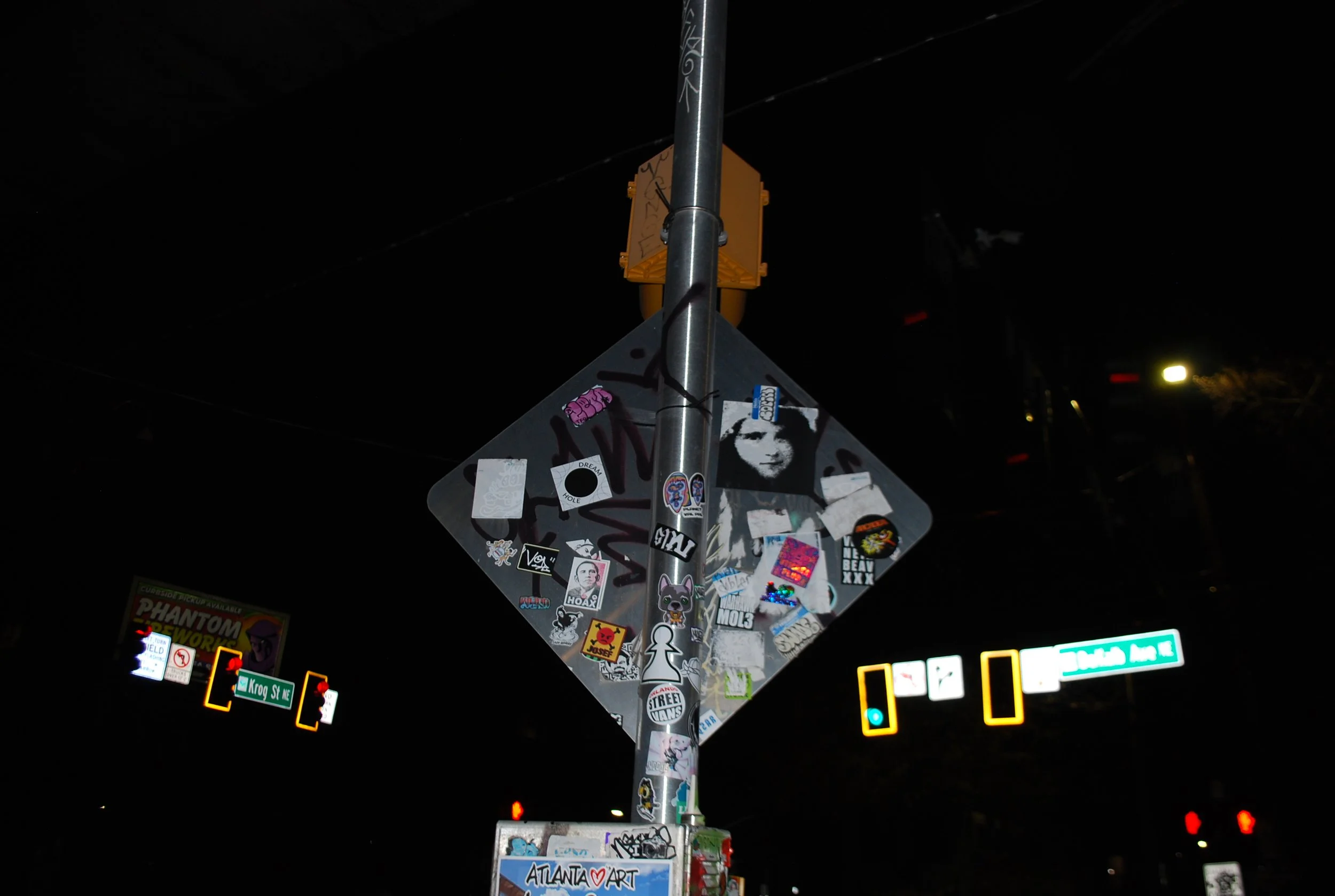 Street pole at night covered with stickers, street signs, and traffic lights. The street signs read 'Krog St NE' and 'Johns Ave'.