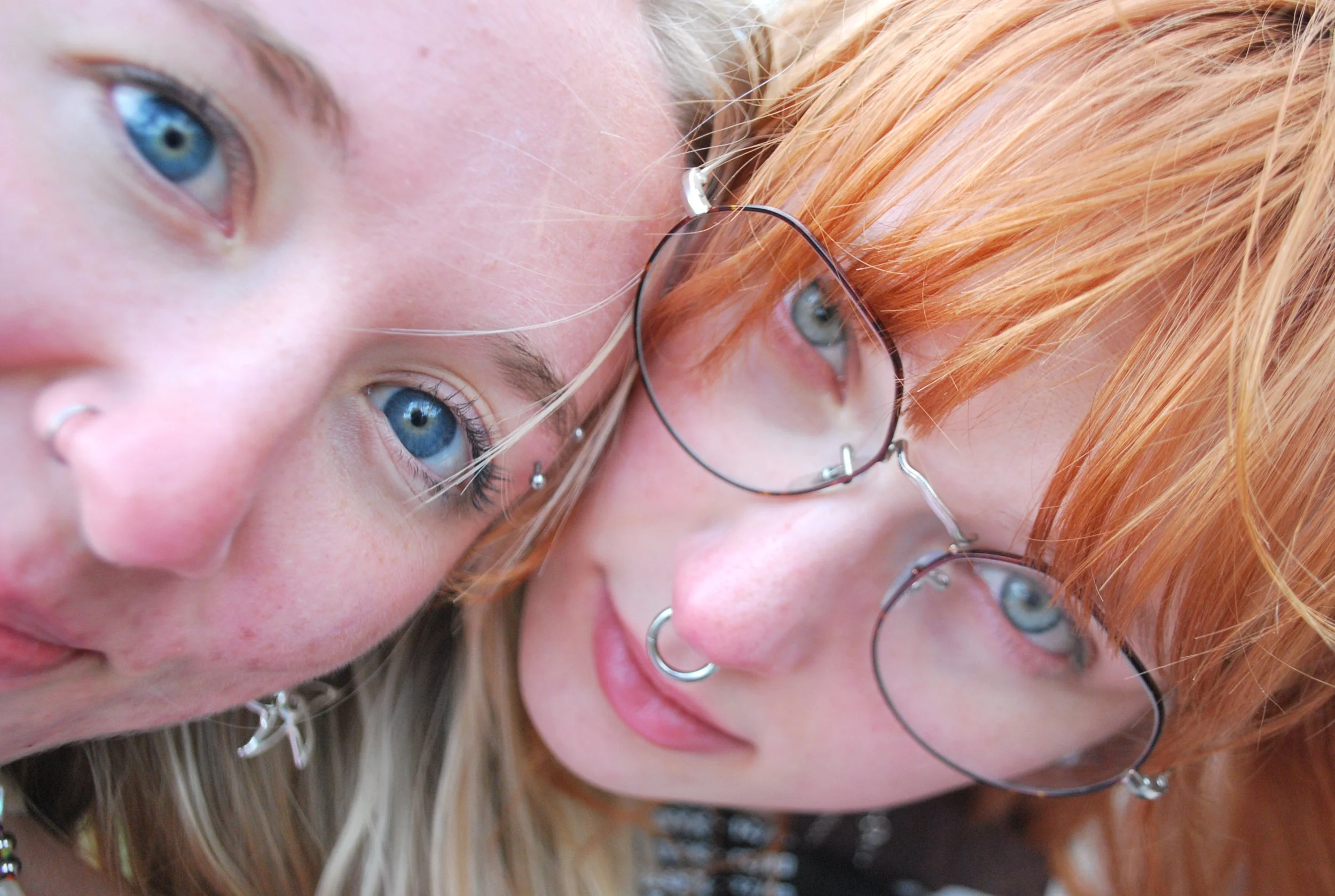 Close-up photo of two young women with blue eyes and piercings, one with orange hair and glasses, the other with blonde hair.