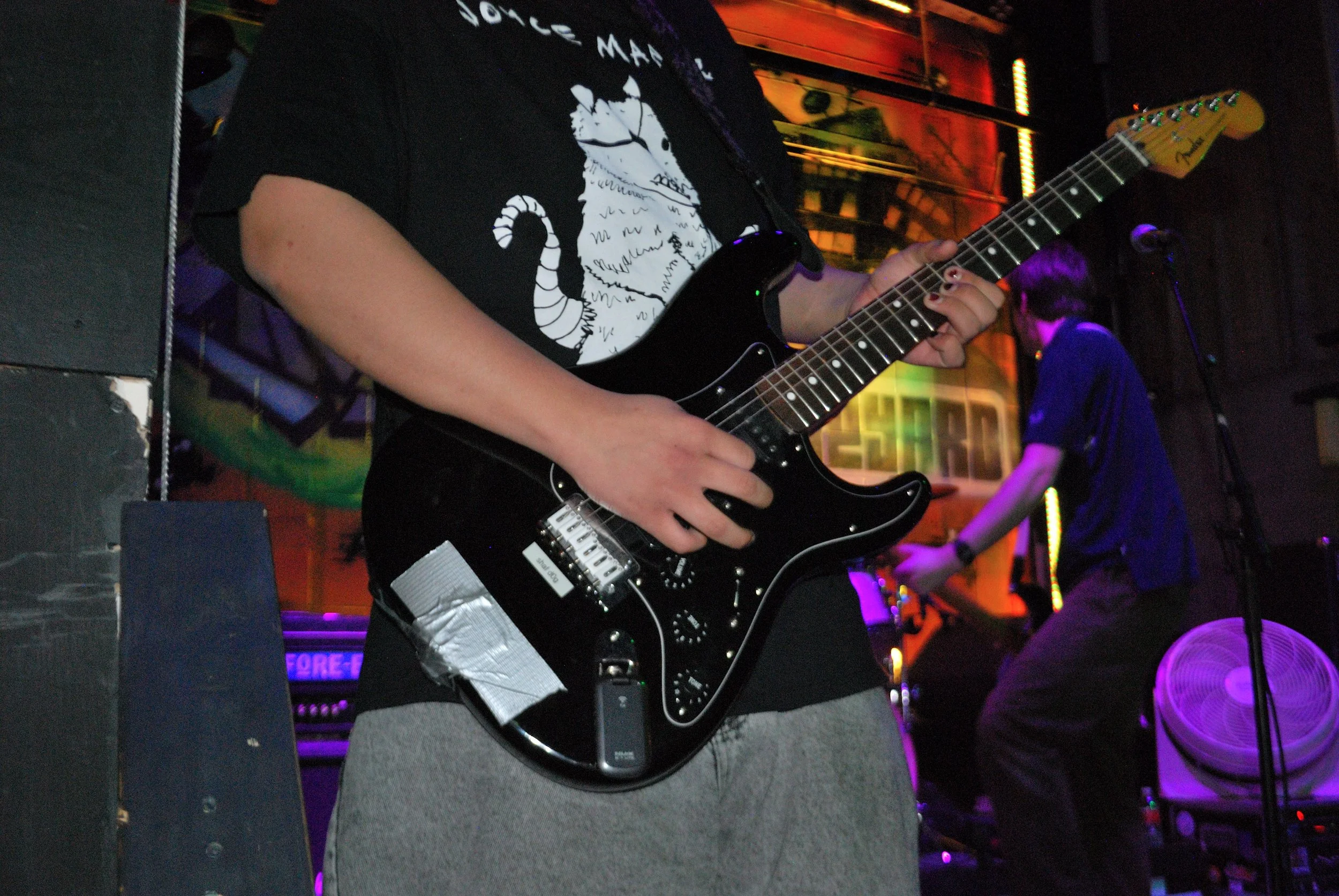 Person playing a black electric guitar on stage at a concert, with others in the background and colorful stage lighting.