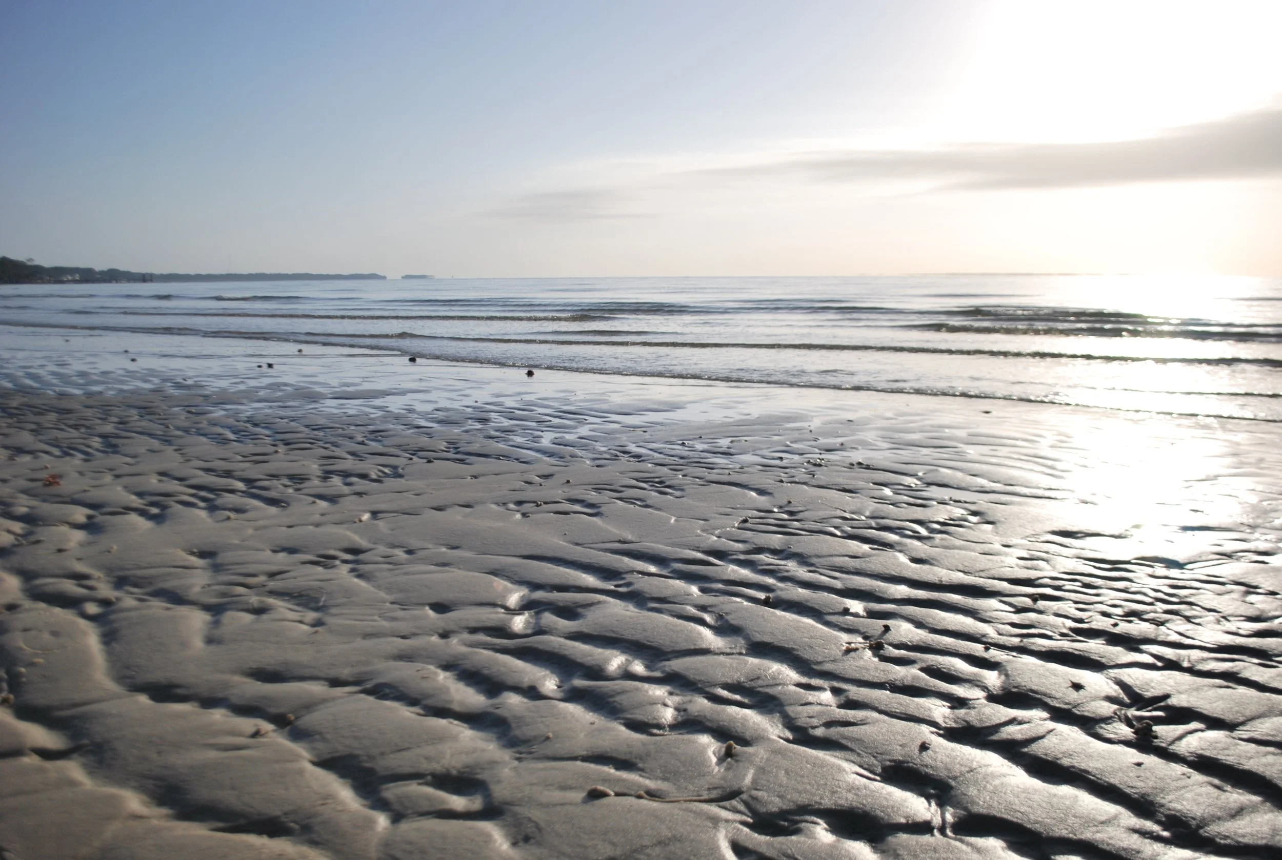 Sunlit beach with wet sand ripples and gentle ocean waves, horizon line, and cloudy sky.
