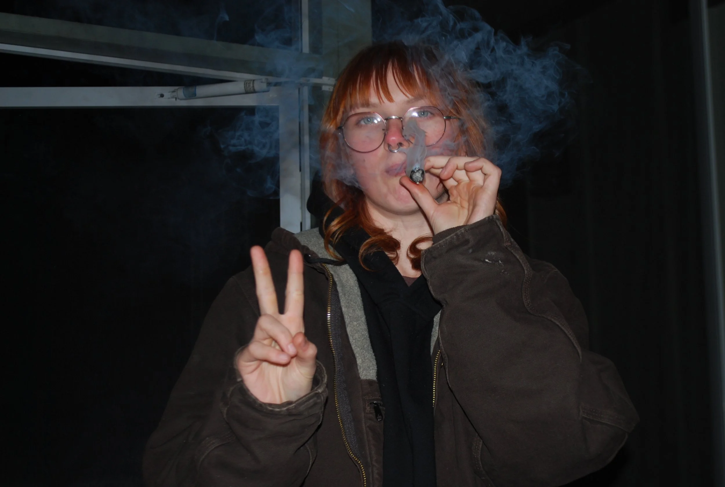 Person with red hair, glasses, and a nose ring smoking a cigarette and making a peace sign with their left hand