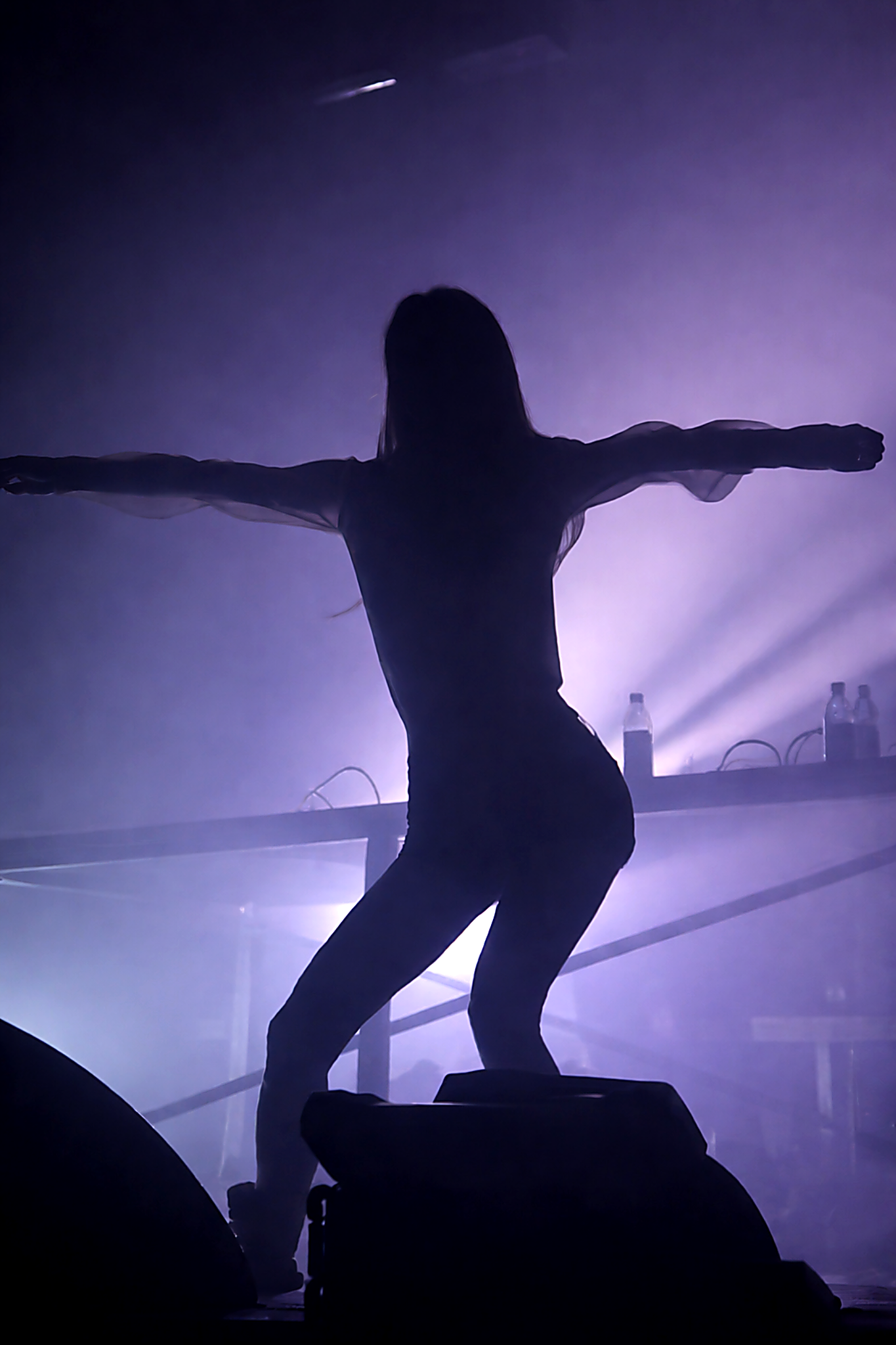 Silhouette of a woman with arms outstretched on stage with purple backlight, DJ equipment, and water bottles behind her.