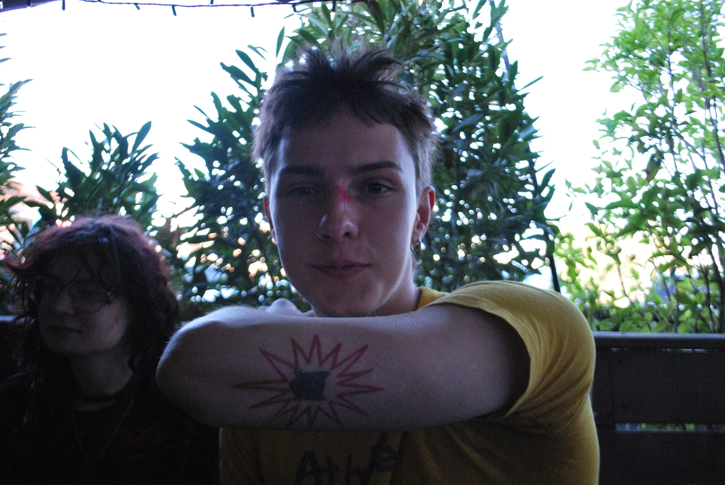 Young man with short hair, nose ring, and tattoos on his arm, sitting outdoors in a shaded area with greenery in the background. Another person with dark, curly hair and glasses is partially visible beside him.