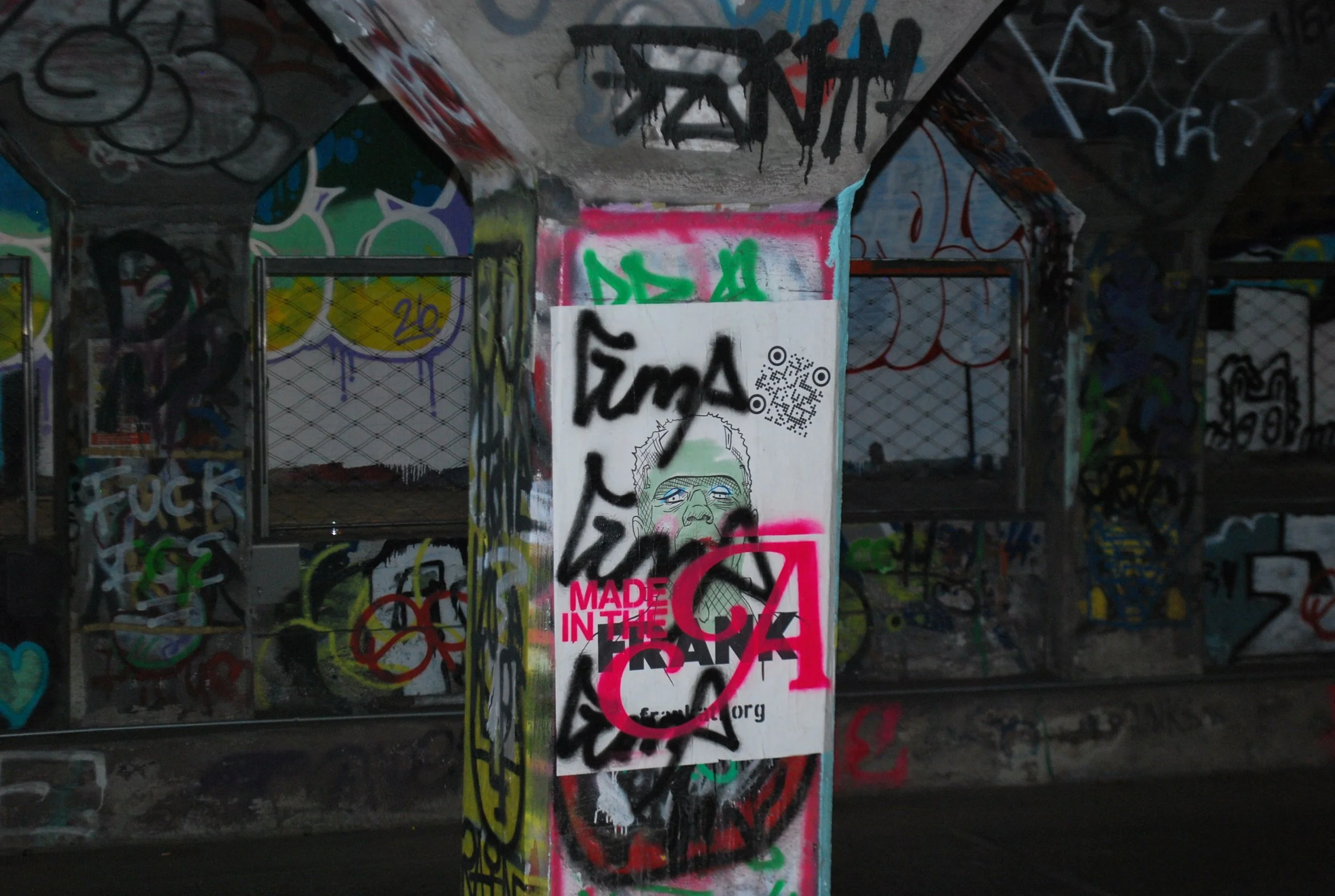 A graffiti-covered urban structure with a central pillar featuring a sticker that says "Made in the Fake" and includes a cartoon face and QR code, surrounded by colorful graffiti on walls and fences.