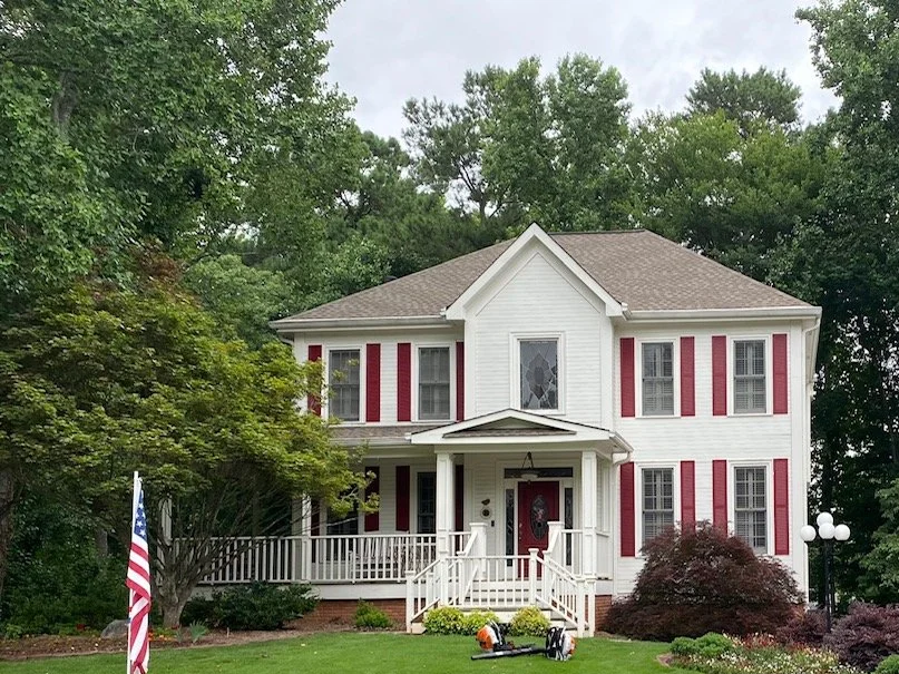 Two-story white house with red shutters and a covered front porch, surrounded by trees and a well-maintained yard.