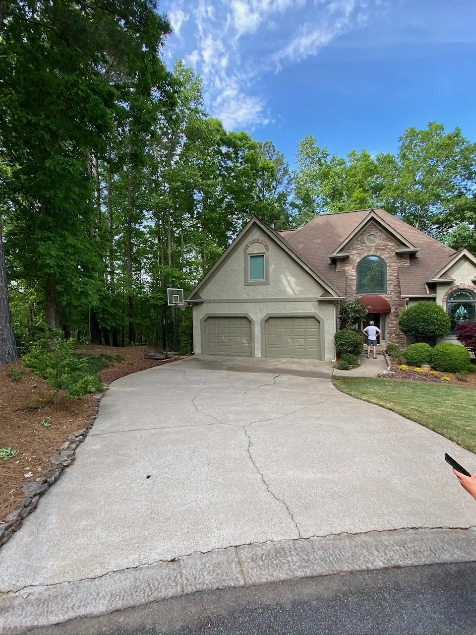 A residential house with a double garage, surrounded by greenery and trees. There is a basketball hoop on the side of the driveway and a person with a hat walking near the entrance. The sky is partly cloudy with blue skies visible.
