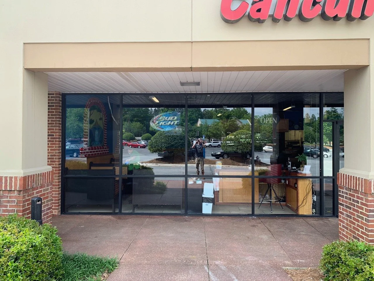 View of a restaurant storefront with large glass windows, a sign that reads 'Canteen,' and a reflection of parking lot and trees outside.