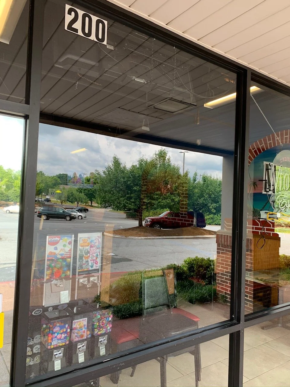 View through a large glass window showing a parking lot with trees and cars outside and vending machines inside a convenience store at 200 block.