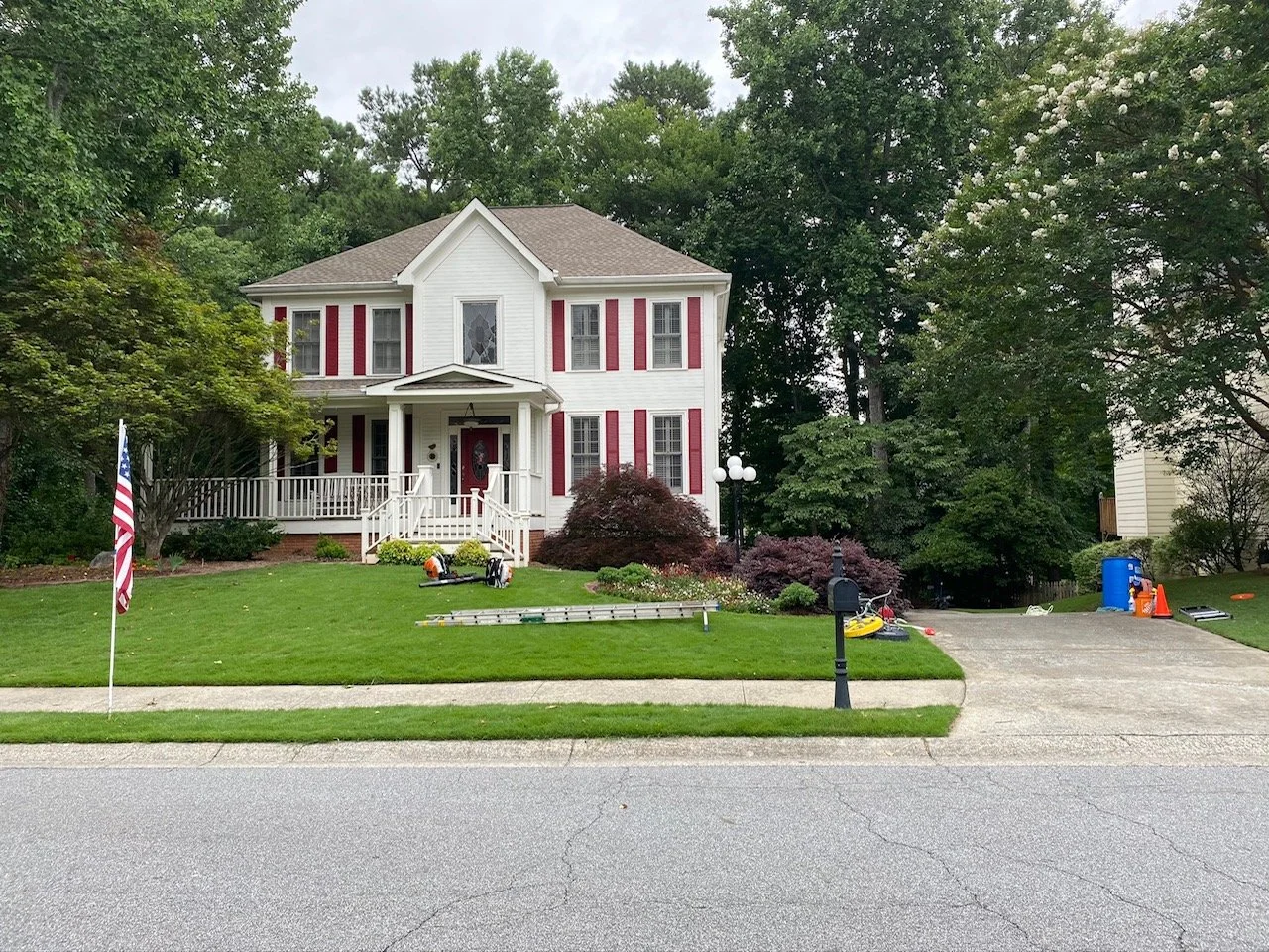 Two-story white house with red shutters, front porch, and wooden railing. Well-maintained lawn with bushes and a tree. Driveway on the right with gardening tools, a kayak, and outdoor cones, and a blue trash bin in the backyard.