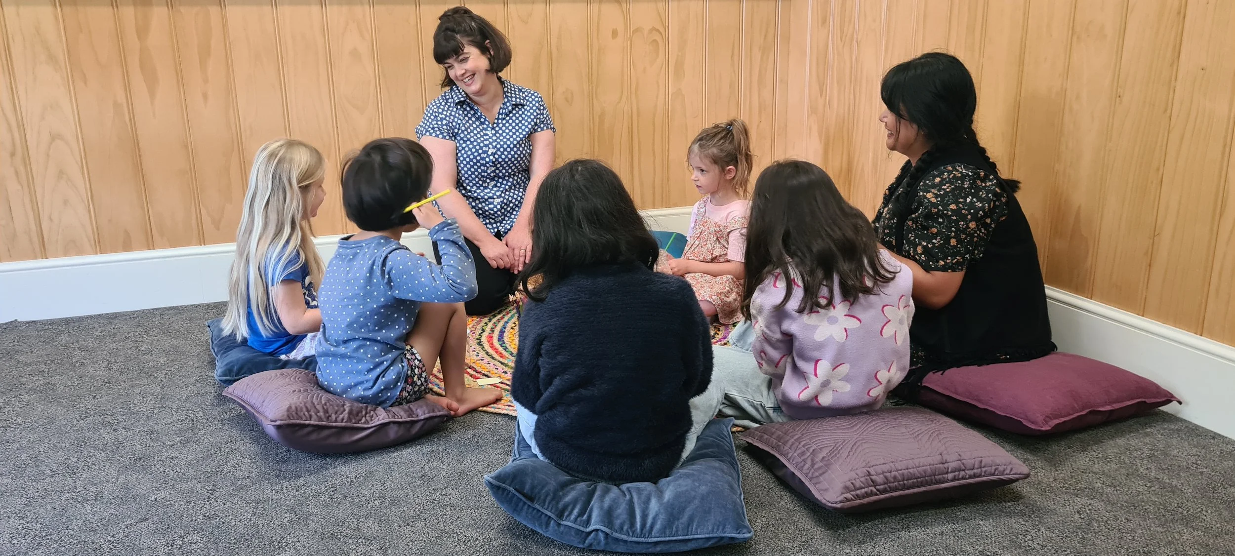 Two women leading a group of children in a story or activity while seated on floor pillows on a carpeted room with wooden panel walls.