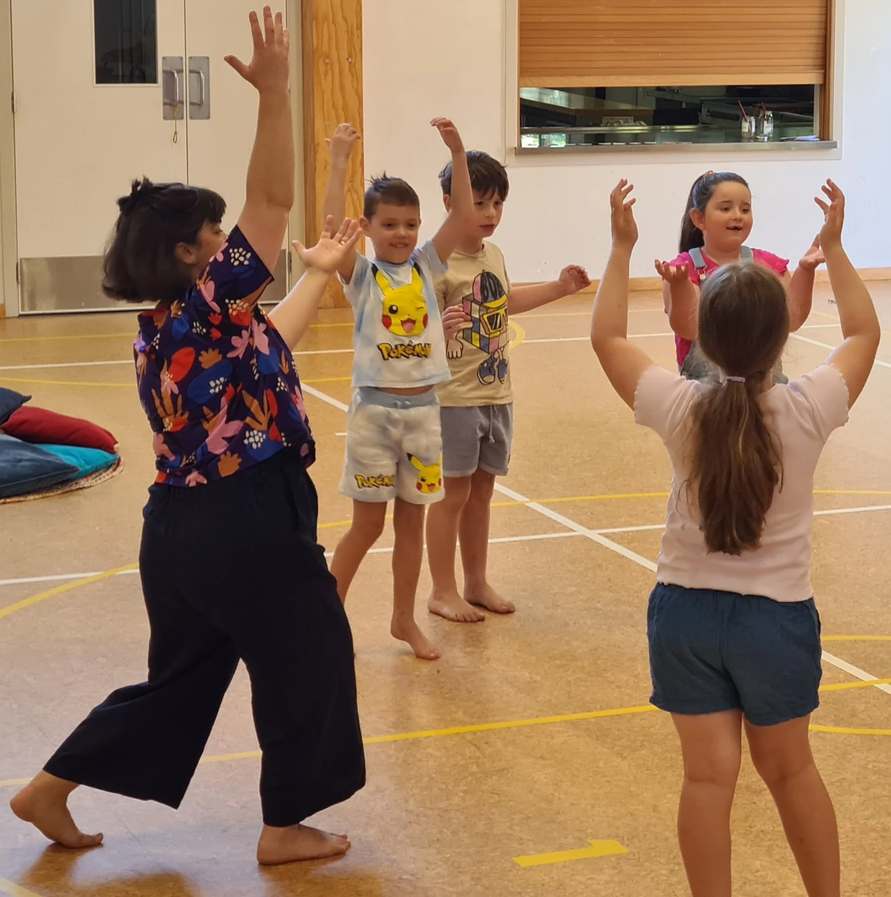 A group of children and a teacher in a gymnasium participating in a balancing activity. The teacher is holding her arm up, and the children are standing on one leg, with arms raised, balancing on the floor.