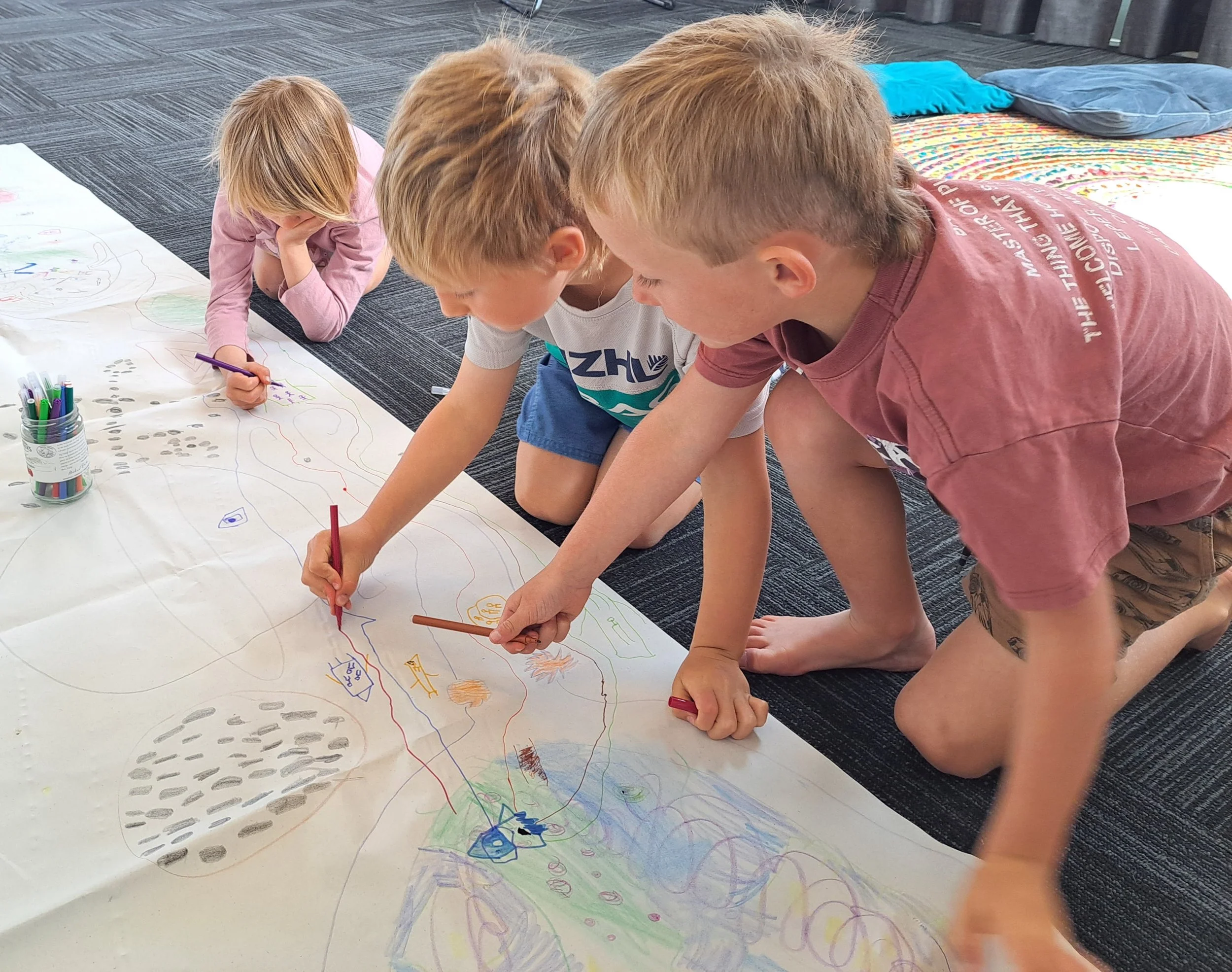 Four children gathered on the floor, drawing and coloring on a large sheet of paper with colorful markers.