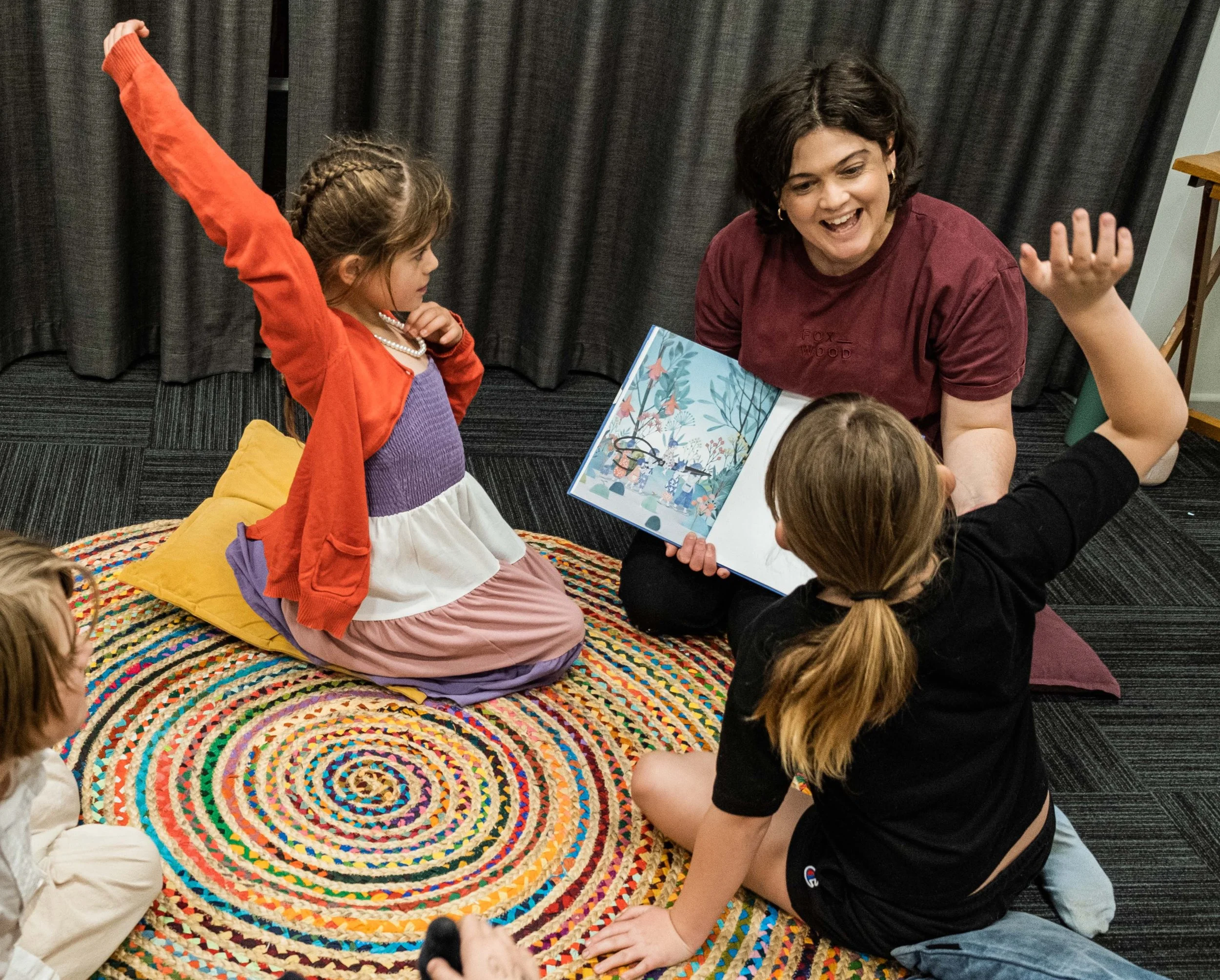 A woman and children sitting on a multicolored spiral rug, engaging with a children’s book, with one girl raising her arm, in a cozy indoor setting with dark curtains in the background.
