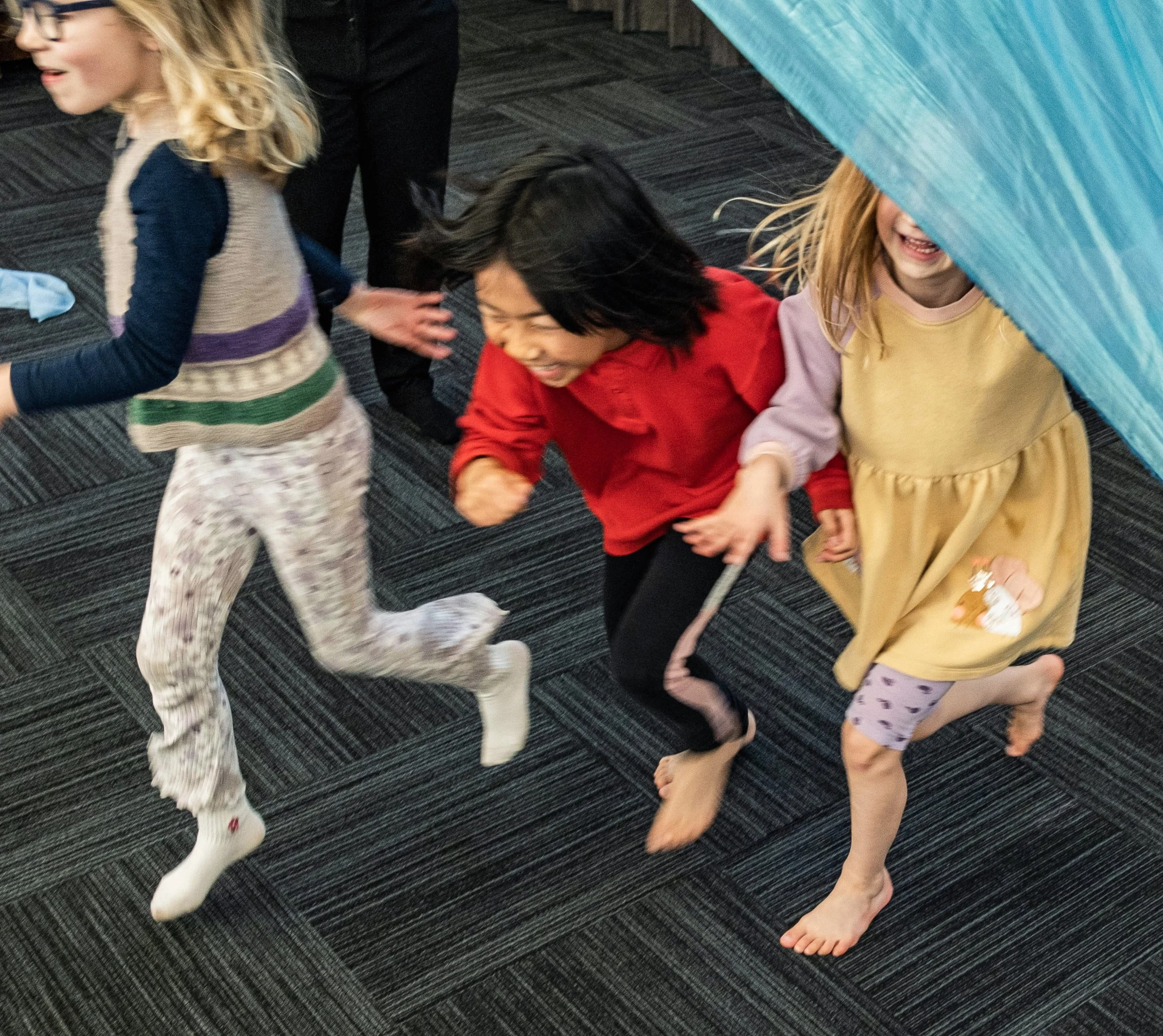 Three children playing on a black carpeted floor; one girl with long blond hair wearing glasses and a navy and beige sweater, another girl with blond hair wearing a yellow dress partially obscured by a blue object, and a girl with black hair wearing a red jacket; all are smiling and barefoot.