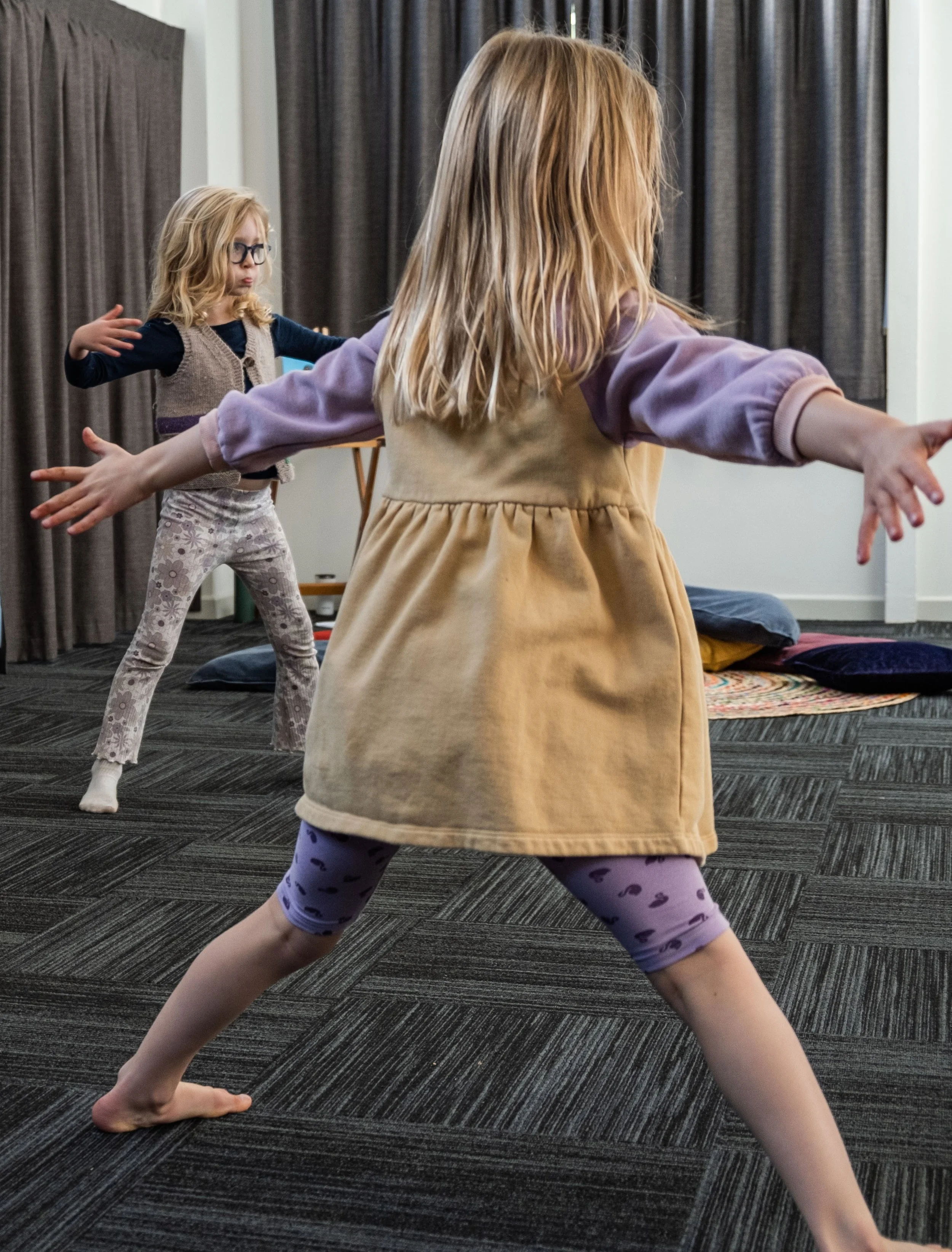Two young girls practicing yoga or dance in a room with dark curtains and a carpeted floor. One girl in the foreground with blonde hair, wearing a beige dress with purple sleeves and purple leggings with black heart patterns. The other girl in the background with blonde hair and glasses, wearing a beige vest over dark top and floral leggings.