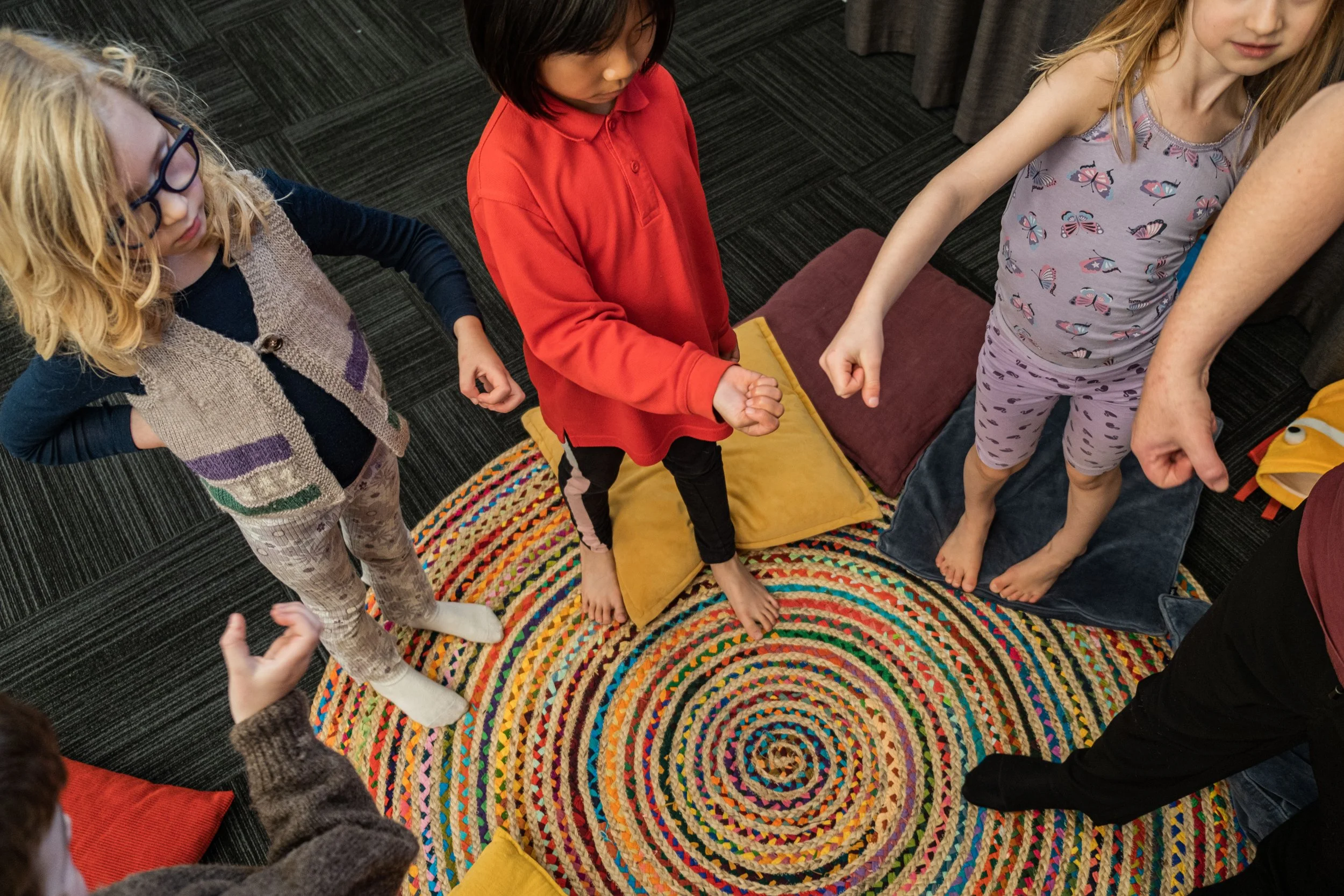 Children standing around on a multicolored spiral rug, with their fists clenched and arms extended downward, in a group activity.