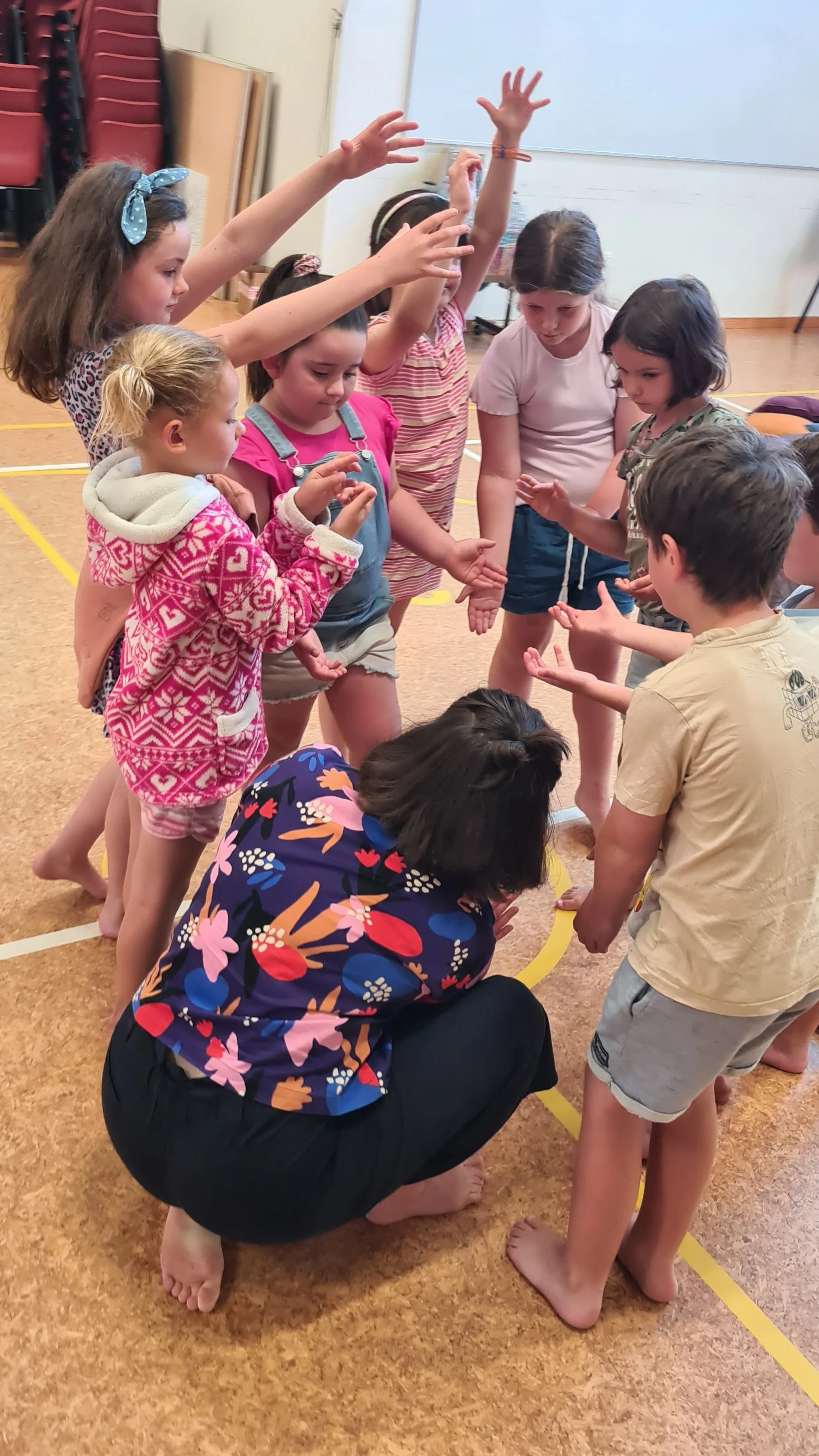 A group of children and an adult are gathered in a circle on a gymnasium floor, playing a game together. The children are reaching toward the center, and some are barefoot. The adult, kneeling, is wearing a colorful, floral shirt.