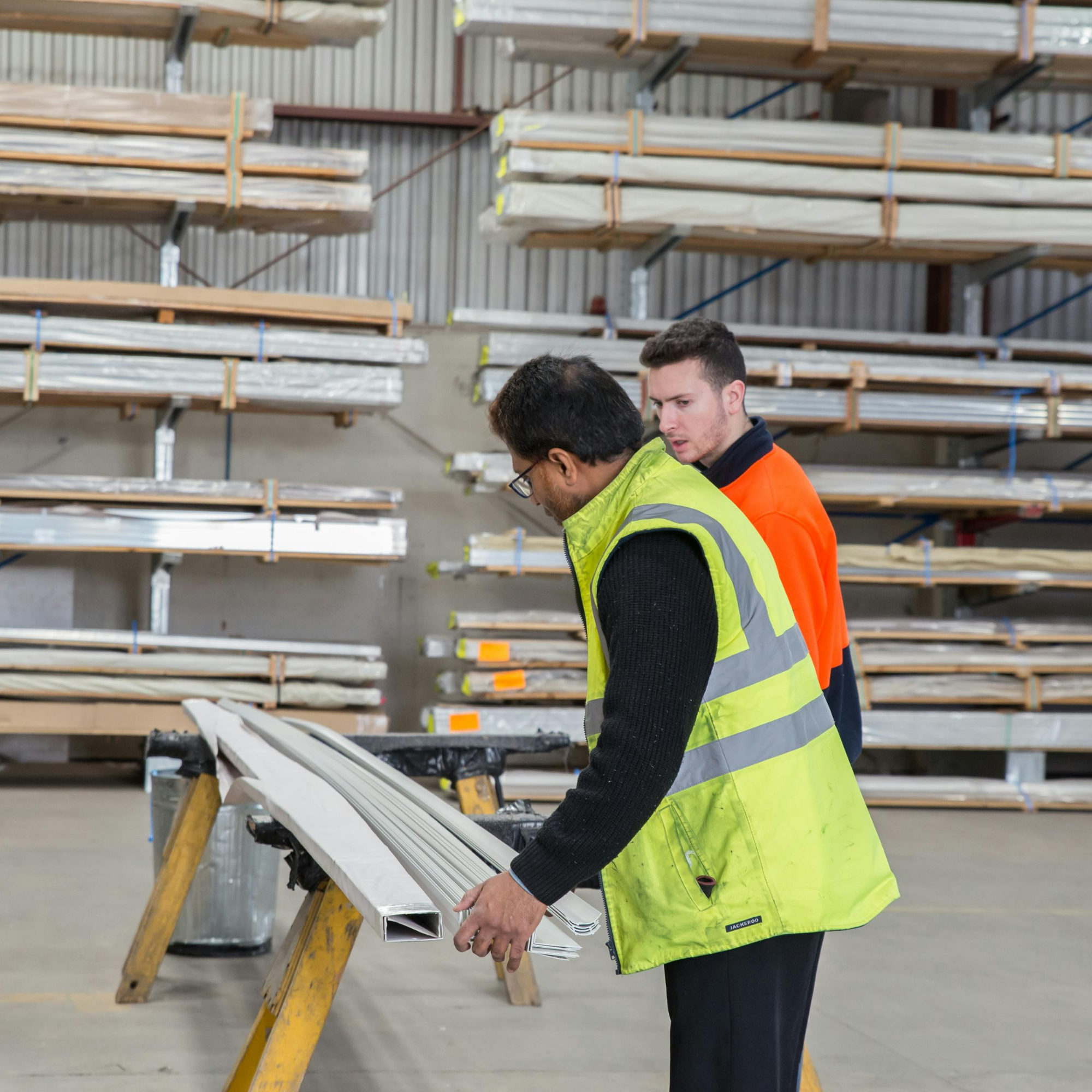 Two workers in safety vests inspecting materials in a warehouse with shelves of metal and wood planks.