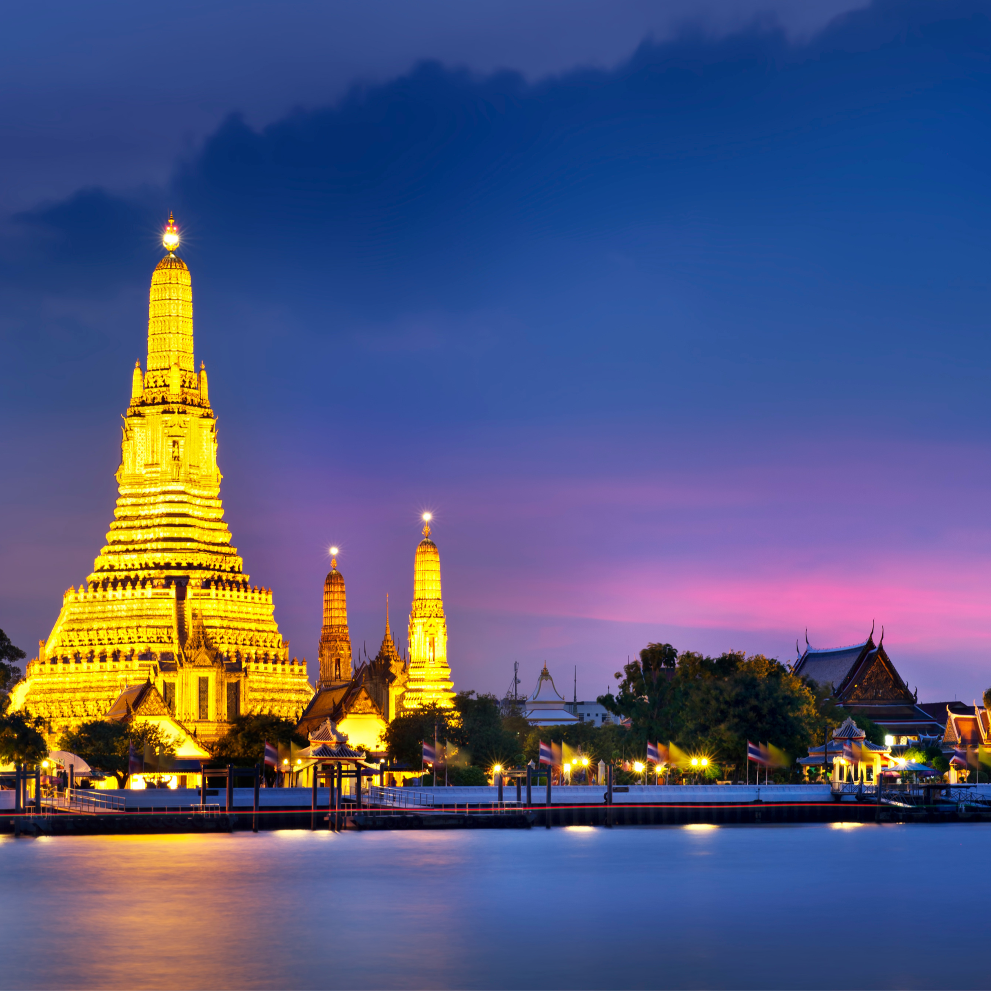 Night view of Wat Arun temple illuminated by yellow lights, reflecting on river with purple and blue sky background.