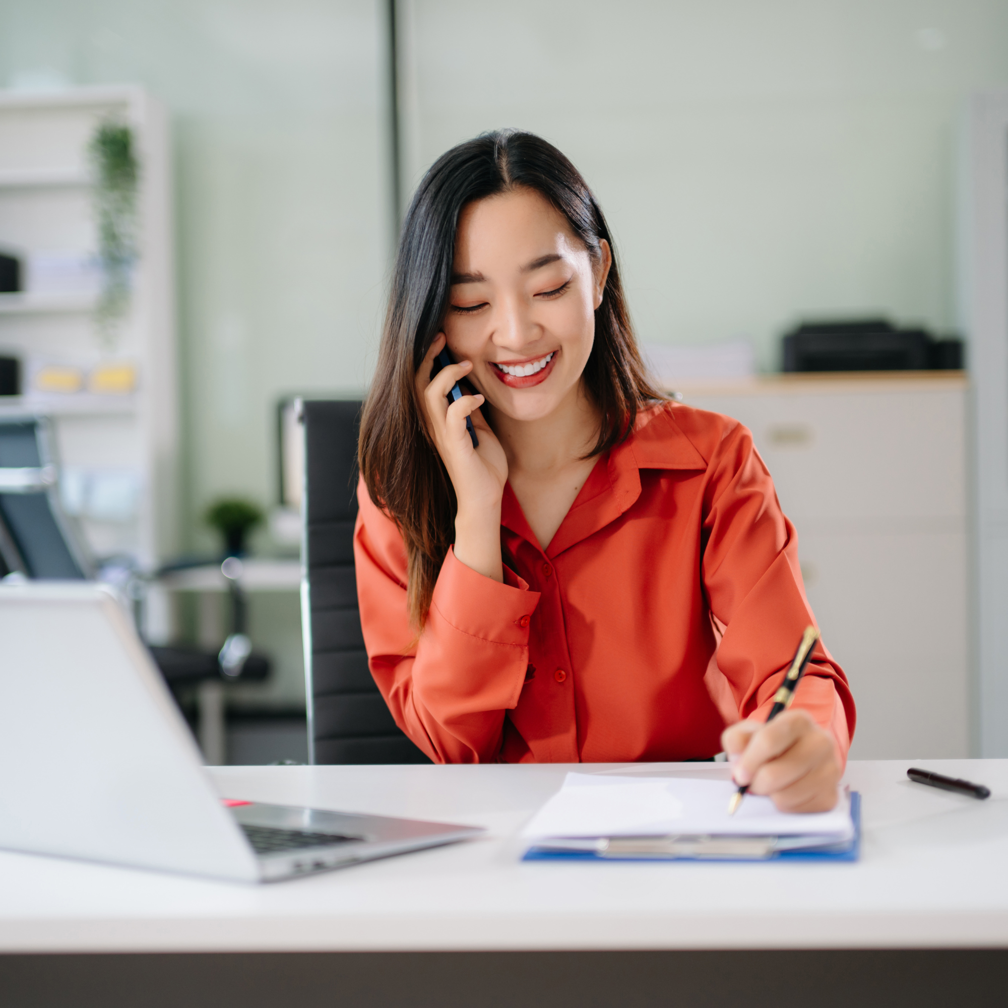 A woman smiling while talking on her cellphone in an office setting, with a desk, laptop, and office supplies in front of her.