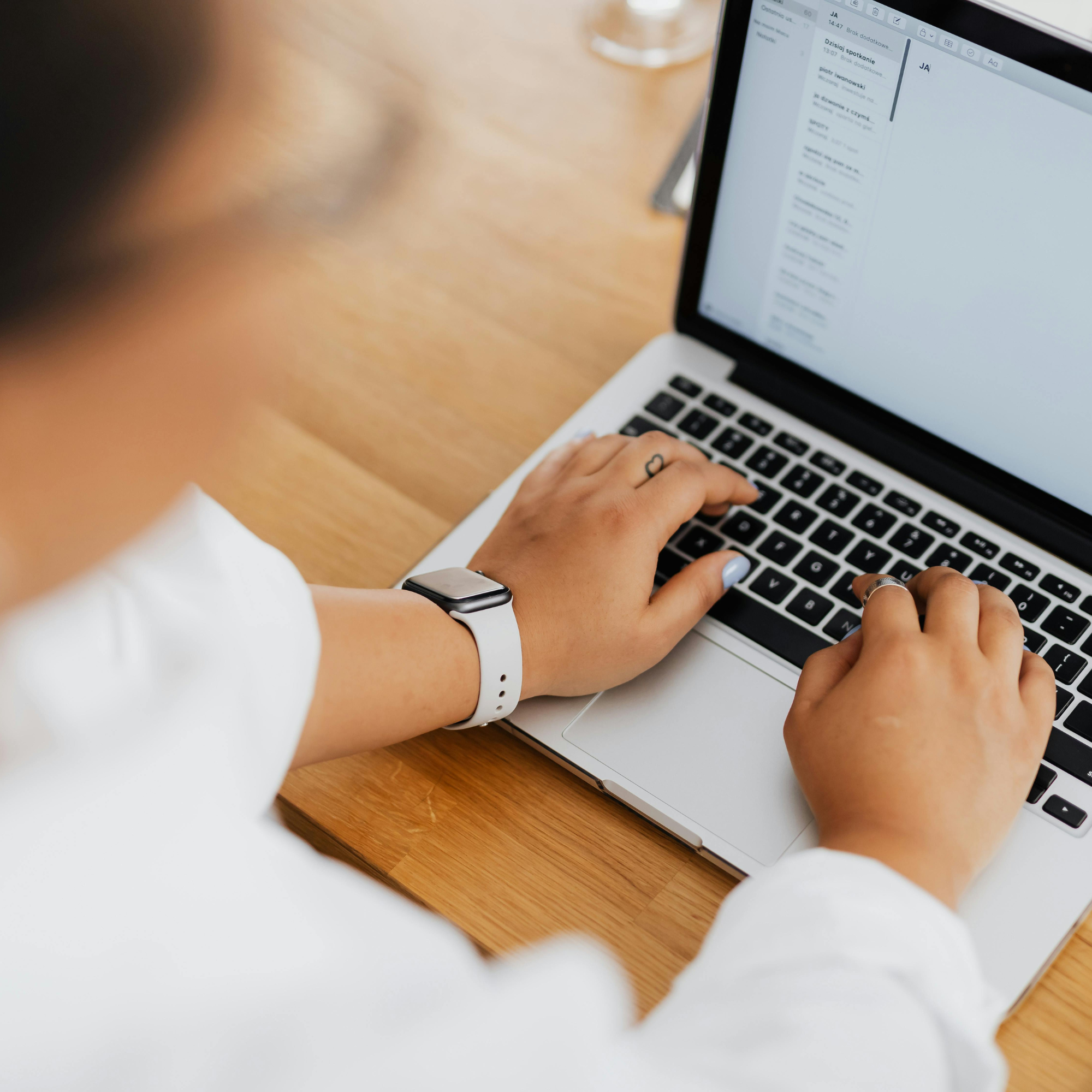 Person using a laptop at a wooden desk with a glass of drink. The person has a wristwatch and a ring, with a short manicure and writing on the laptop screen.
