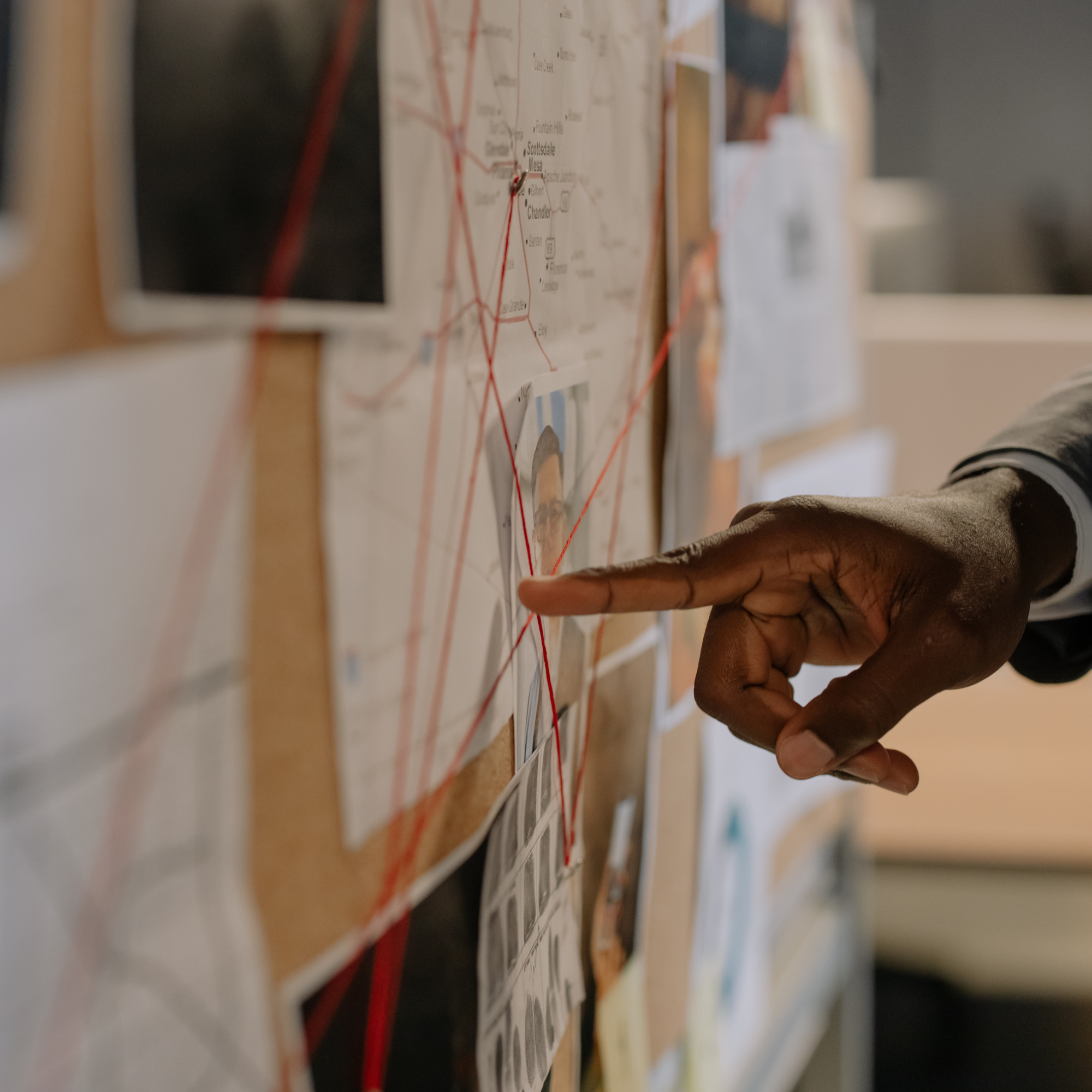 Close-up of a bulletin board with various papers, notes, and images pinned or attached, including a map with red lines indicating routes or connections, and a person's hand pointing or attaching something.