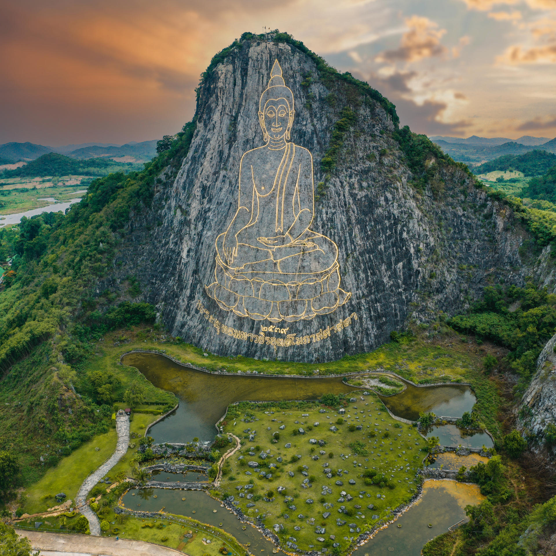 A large mountain with an outline of a sitting Buddha carved into the rock face, overlooking a landscape of small ponds and greenery, during sunset.