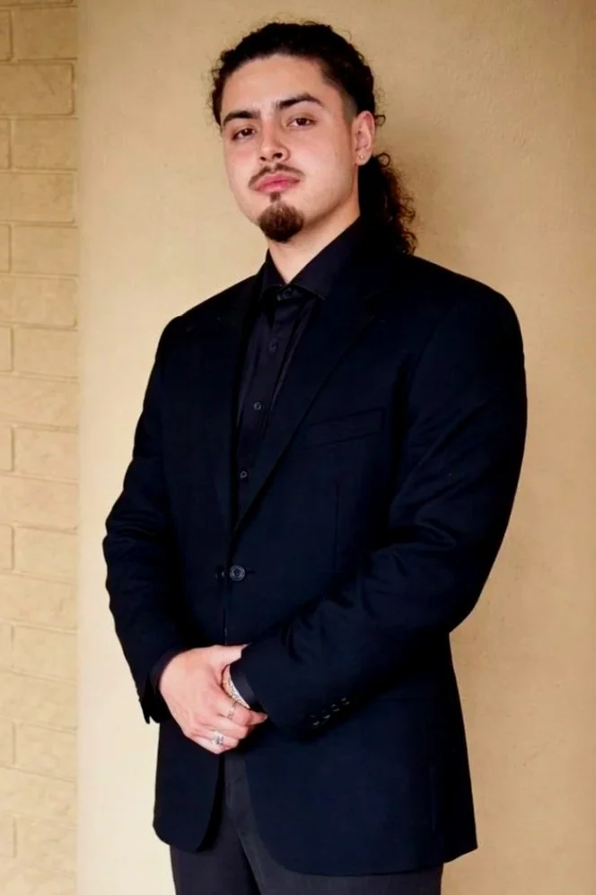 A young man with dark, curly hair tied back, wearing a black suit and shirt, standing against a beige background with a brick wall on the left.
