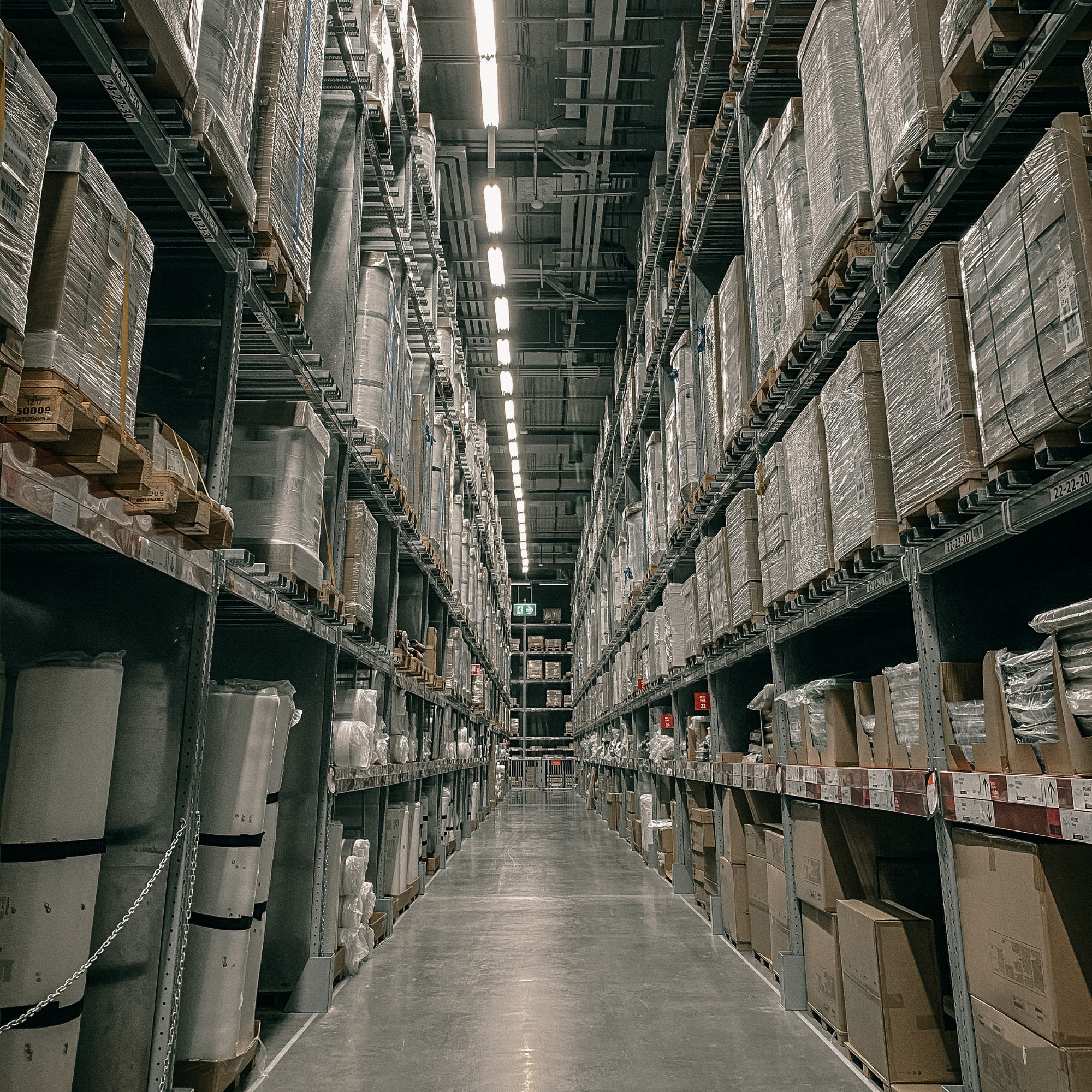 Warehouse aisle with tall metal shelves stocked with boxed and wrapped items, illuminated by overhead fluorescent lighting.