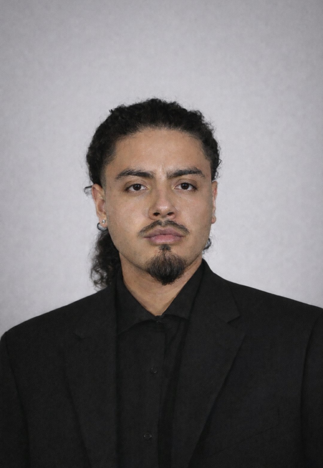 A man with curly black hair, facial hair, and earrings, dressed in a black suit and shirt, standing against a plain light gray background.