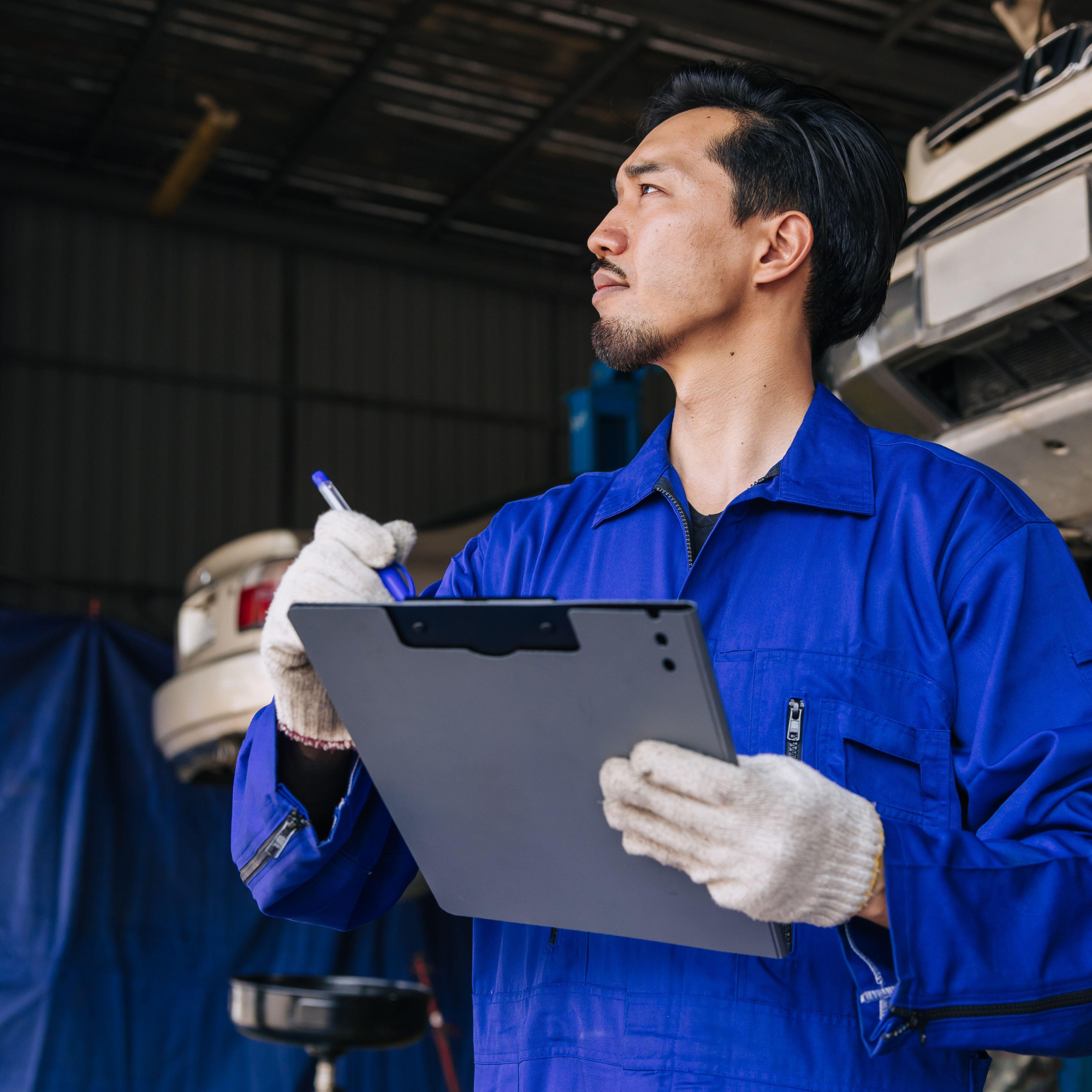 A man with black hair and a goatee wearing a blue work jacket is holding a tablet in one glove and a pen in the other, standing in what appears to be an industrial or workshop setting with vehicles in the background.