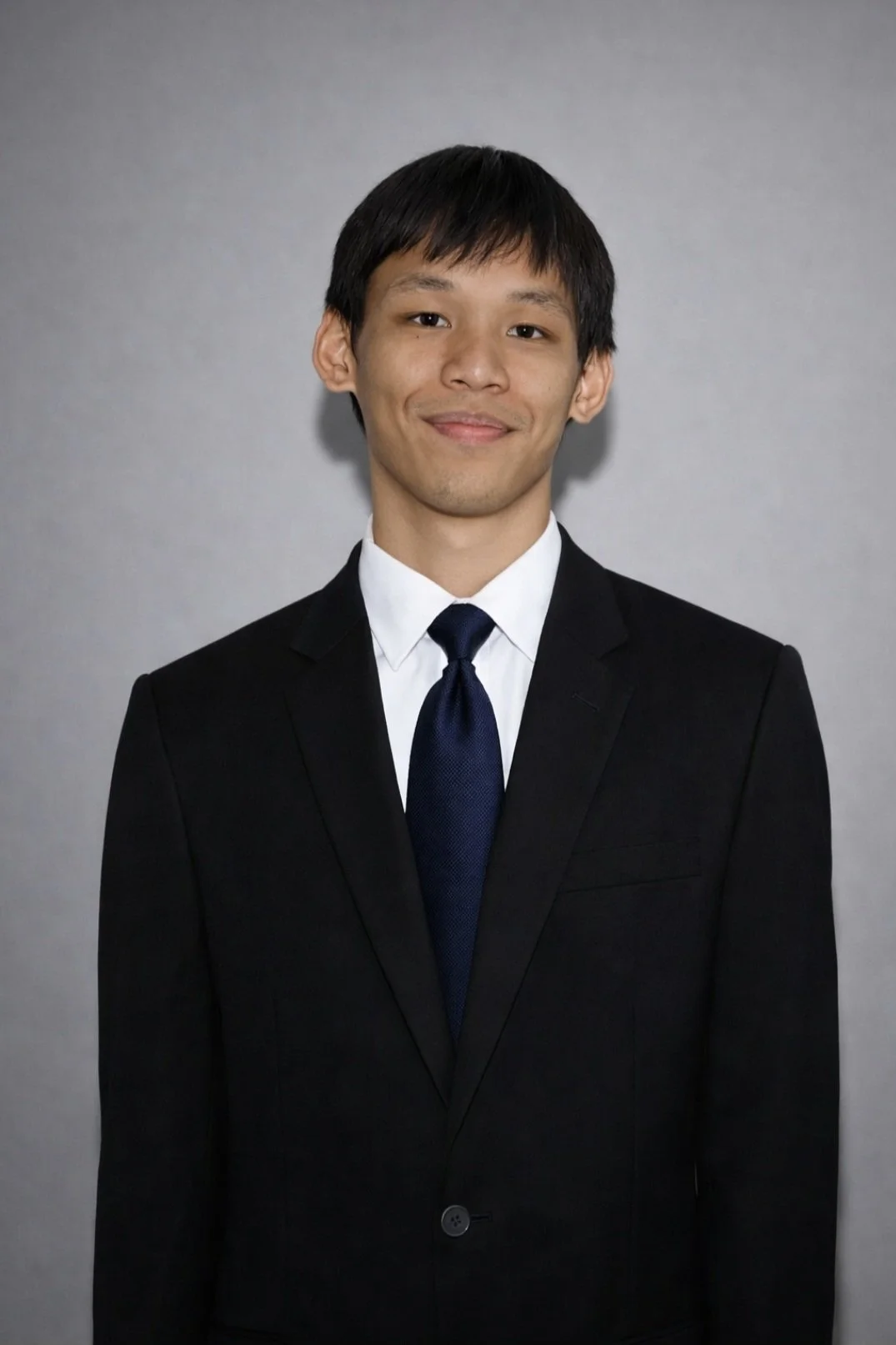 A young man in a black suit, white dress shirt, and dark tie, standing against a plain gray background.
