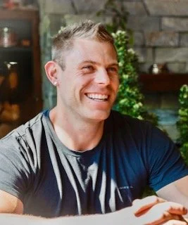 A smiling man with short gray hair, wearing a black and gray t-shirt, sitting indoors near a decorated Christmas tree.