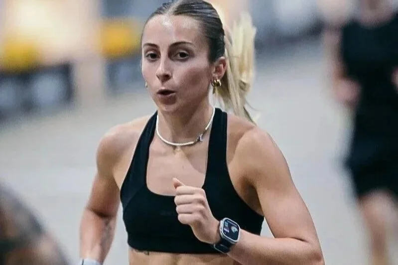 A woman running in a race, wearing a black sports bra, a smartwatch, and a necklace.