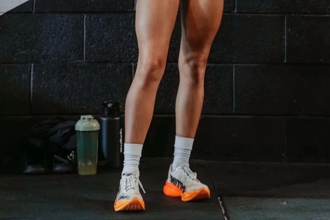 Close-up of a person’s athletic legs, wearing white socks and running shoes, in a gym setting with water bottles and gym bag in the background.
