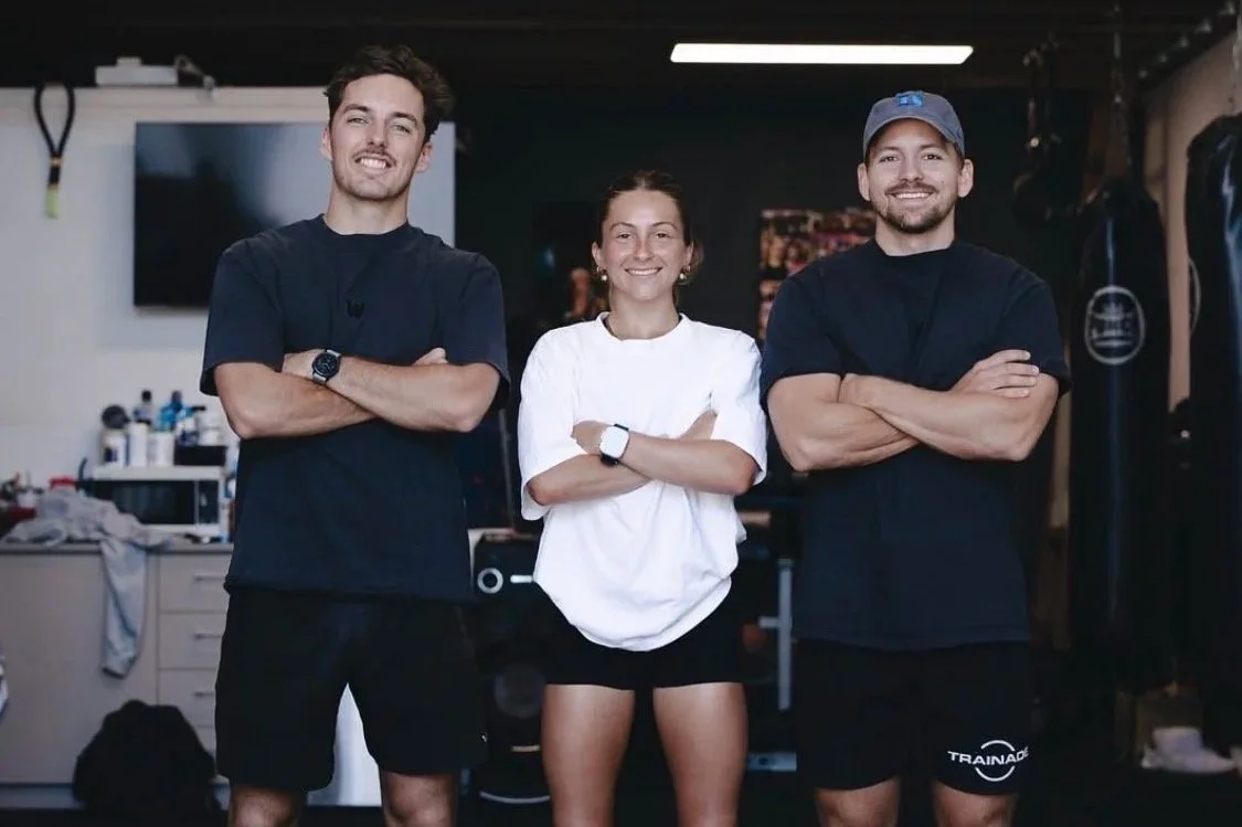 Three young adults, two men and one woman, standing inside a gym or fitness studio with arms crossed and smiling at the camera.