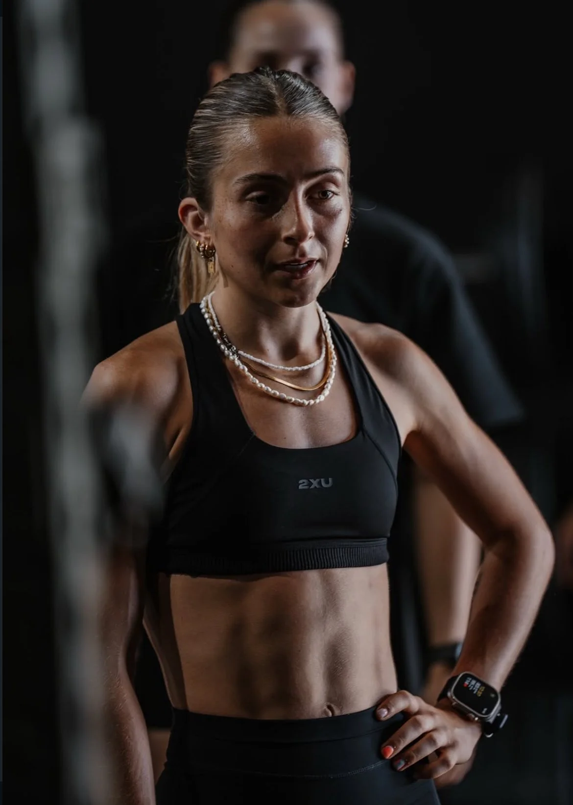A woman wearing athletic clothing and jewelry, standing in a gym, looking contemplative, with a blurred dark background and a person behind her.