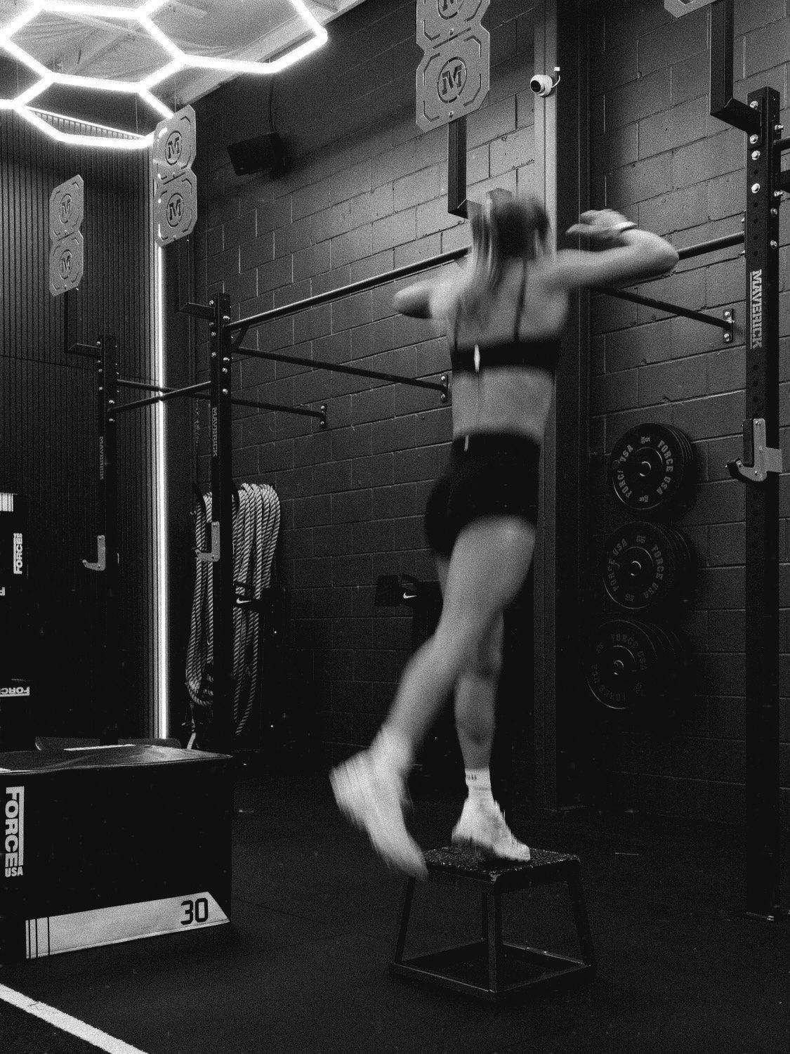 A woman in athletic clothing performs a box jump exercise in a gym, jumping onto a small box with her arms raised.